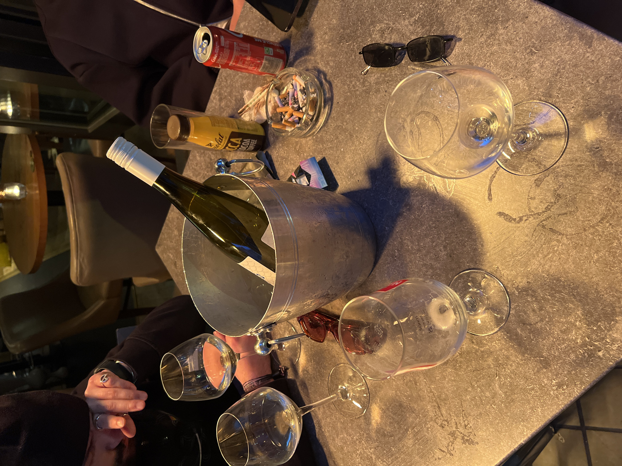 The image depicts an outdoor table setting, likely in London, United Kingdom, due to the style of the setting and objects. The table has a grey, textured surface. At the center of the table is a silver ice bucket containing a bottle of wine. Surrounding the ice bucket are three empty wine glasses. Another empty wine glass and a pair of sunglasses are to the right of the bucket. To the left of the wine bucket is someone's hand with a ring on their finger. In the top left corner, there is a person's sleeve and a can of Coca-Cola. Next to the can is a clear ashtray filled with cigarette butts, straws, and candy wrappers. Next to the ashtray is a bottle of ICA Cocoa Coffee. The environment appears to be a restaurant or bar patio, with chairs and tables visible in the background. The lighting suggests it is evening.