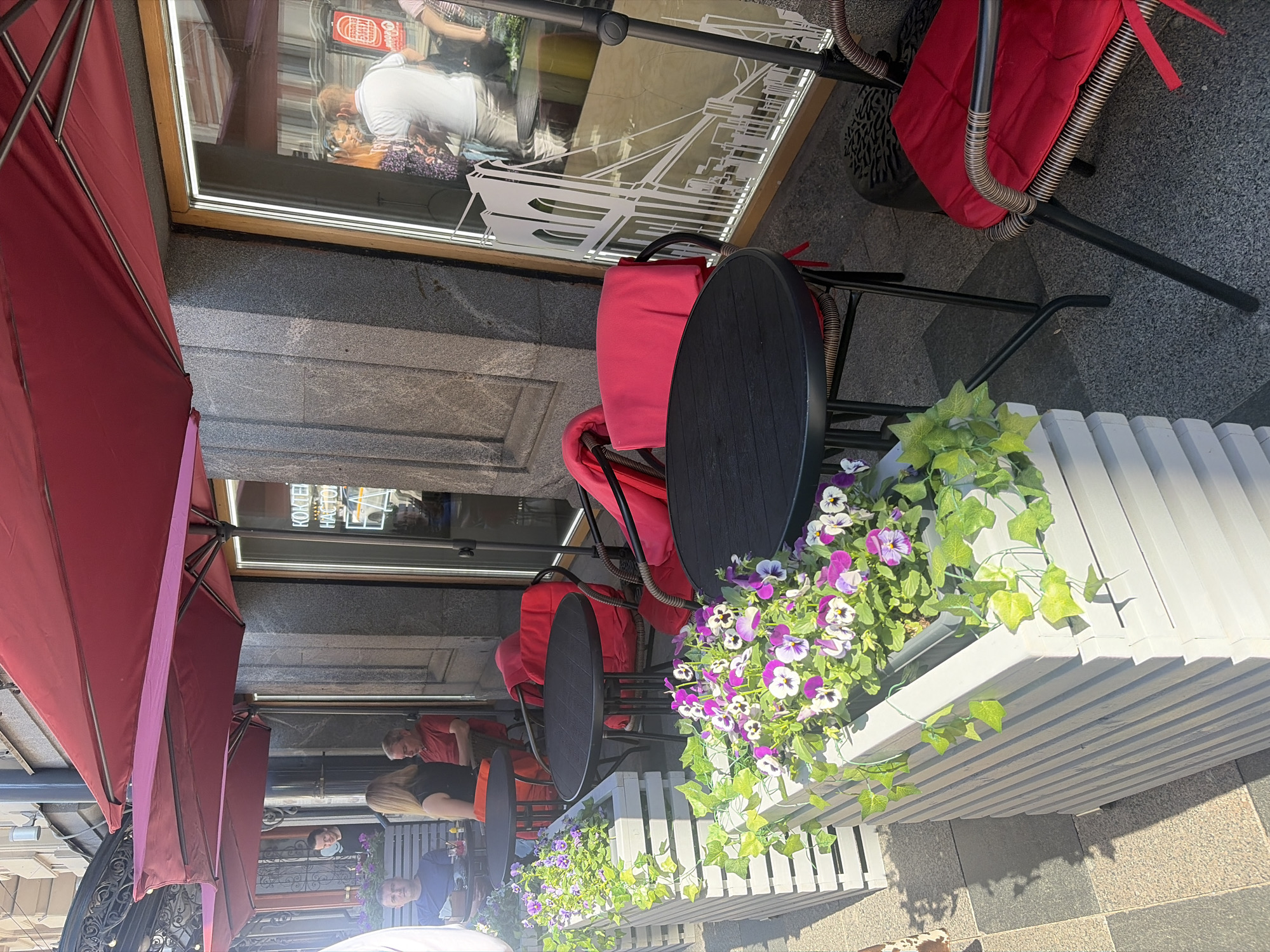 An exterior shot shows a cafe with outdoor seating. Red umbrellas provide shade over a gray stone facade and a sidewalk. There are several black tables and chairs with red cushions. A planter box filled with green ivy and purple and white pansies is in the foreground. Three people are visible at a table on the left, and reflections in the window show more people inside the cafe. The scene is illuminated by sunlight.