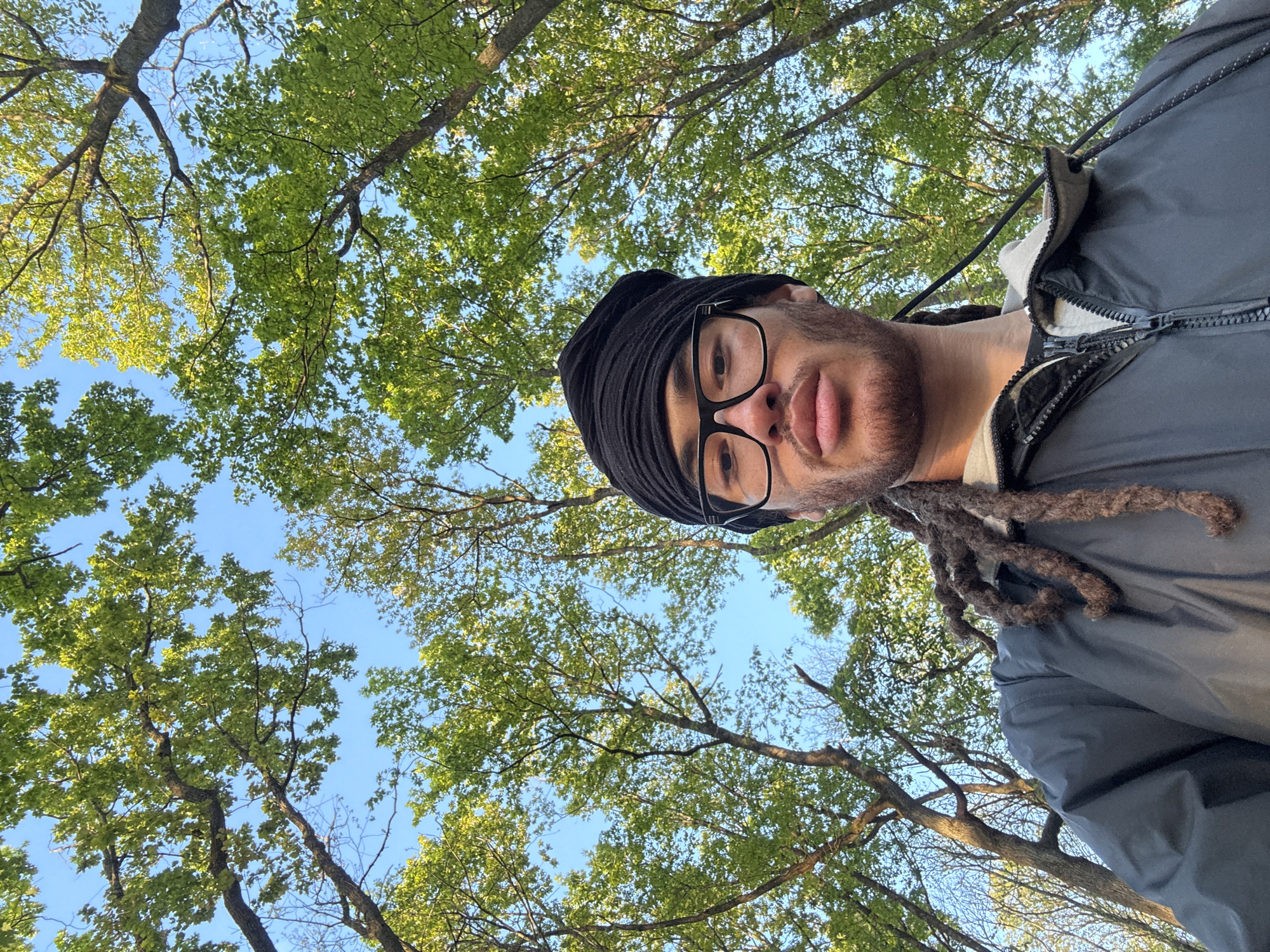 This low-angle outdoor selfie, taken in Bures-sur-Yvette, France, captures an adult male looking directly at the camera. The subject has dark skin, a short beard, and dreadlocks visible near his right shoulder. He wears rectangular-framed glasses, a dark head covering or turban, and a dark grey or black zip-up jacket over a lighter colored garment. His expression is neutral.
The perspective of the shot is from below, looking upwards. Above and behind the man, a clear, bright blue sky is visible, interspersed with the branches and lush, vibrant green leaves of several trees. The leaves appear full and healthy, suggesting a season of late spring or summer. Bright sunlight illuminates the scene, highlighting the foliage and the right side of the man's face, indicating a sunny day. The image is oriented such that the man's head is on the right side of the frame, giving the impression of him looking down or the camera being tilted and rotated significantly upwards. There is no visible text in the image.