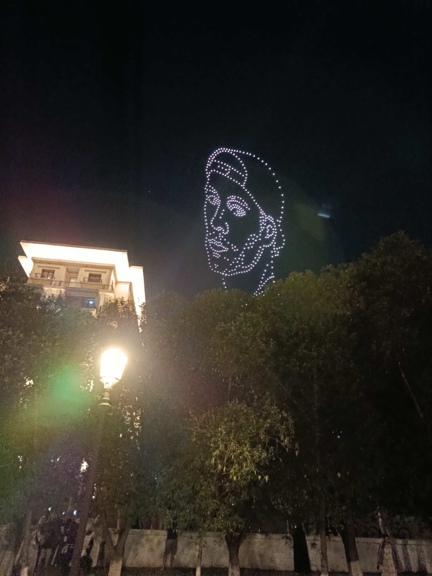 An aerial drone light show forms the outline of a person's face in the night sky above a cityscape. The face is composed of numerous white lights arranged in a dotted pattern, resembling a wireframe. Below the drone display, a modern building with illuminated windows stands behind a canopy of dark trees. In the foreground, a street lamp with a bright, glowing bulb illuminates the area, casting a greenish hue. The ground level appears to be a park or walkway, with several indistinct figures and structures visible among the trees. The overall scene captures a festive or celebratory nighttime event in a city environment, with the drone display being the central visual focus.