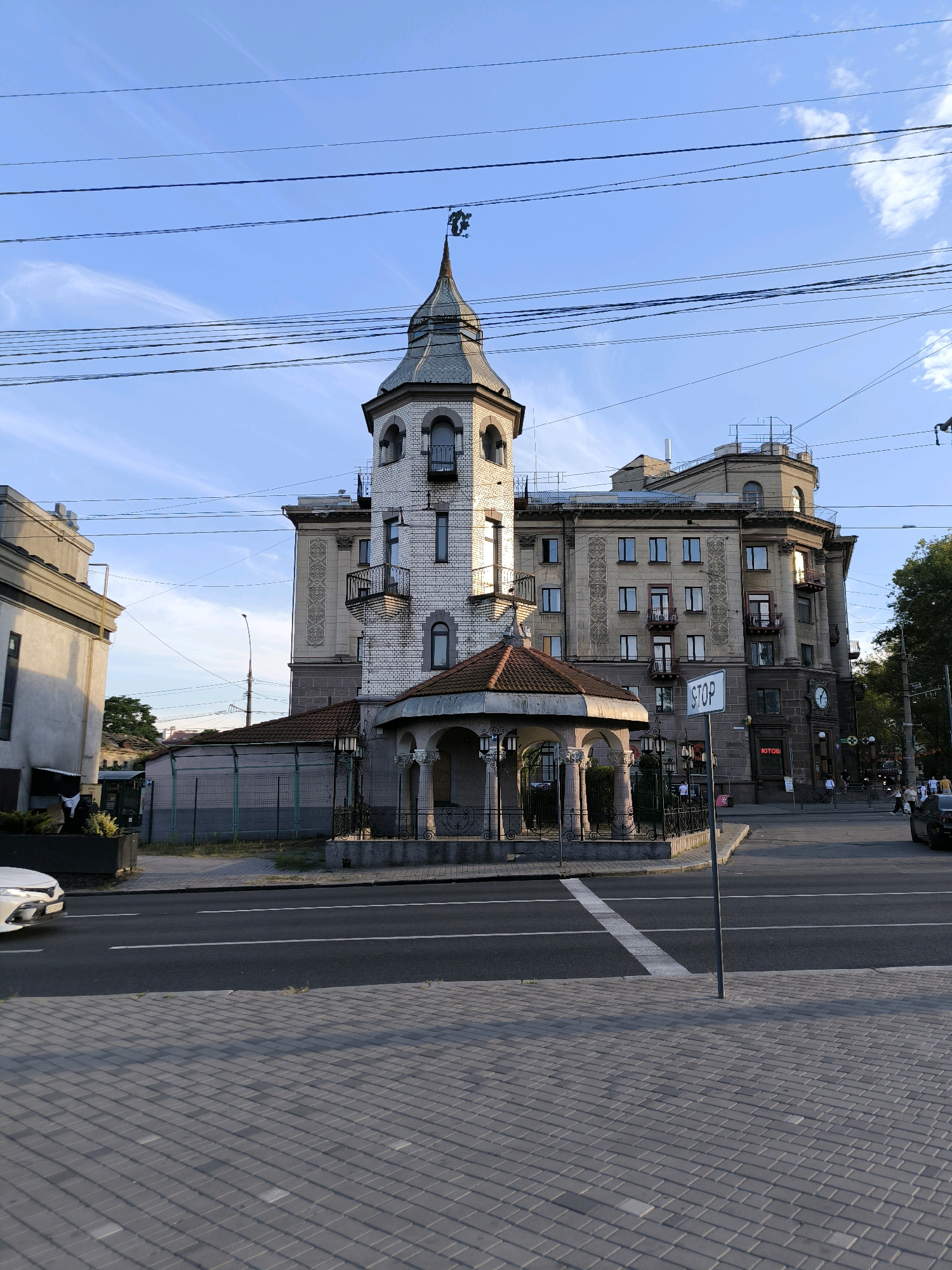This image captures an urban scene in Mykolaiv, Ukraine, featuring a distinctive building with a turret. The building appears to be a multi-story structure with architectural details such as balconies and ornate window frames. A small, covered gazebo or pavilion is situated in front of the main building. A road runs in front of the building, with a pedestrian crossing marked by white lines. Pavement stones make up the sidewalk. Overhead, power lines crisscross the sky, which is a clear blue with wispy clouds. A street sign reading "STOP" is visible near the road. Several people are present on the sidewalk and near the road, some appearing to be pedestrians. Vehicles are also visible on the road. The overall impression is a street-level view of a city environment with a unique architectural focal point.