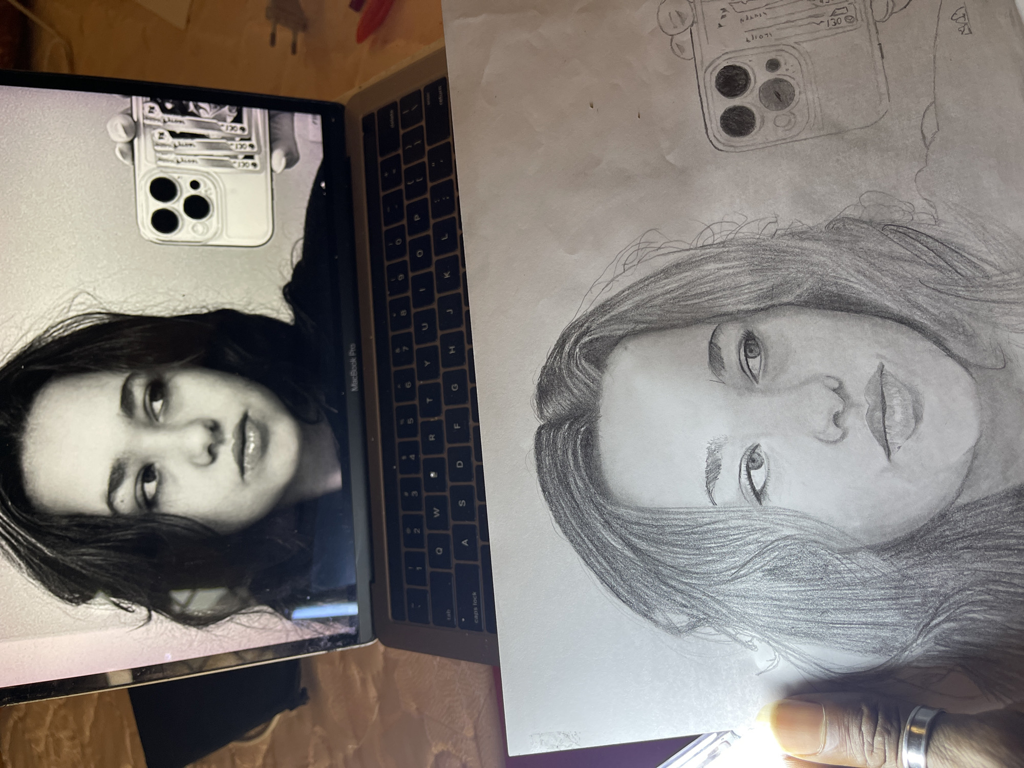 The image shows a laptop with a picture of a young woman on the screen, next to a sketch of the same woman. The laptop is open, displaying the woman's face, and it's placed on a surface near a sheet of paper with the sketched portrait. The sketch includes the woman's face and a representation of the phone case she is holding in the photo. The keyboard of the laptop is visible, and there are some other objects scattered around the edges of the frame, indicating an informal, personal setting. There is no information that indicates a city or location in Nigeria.