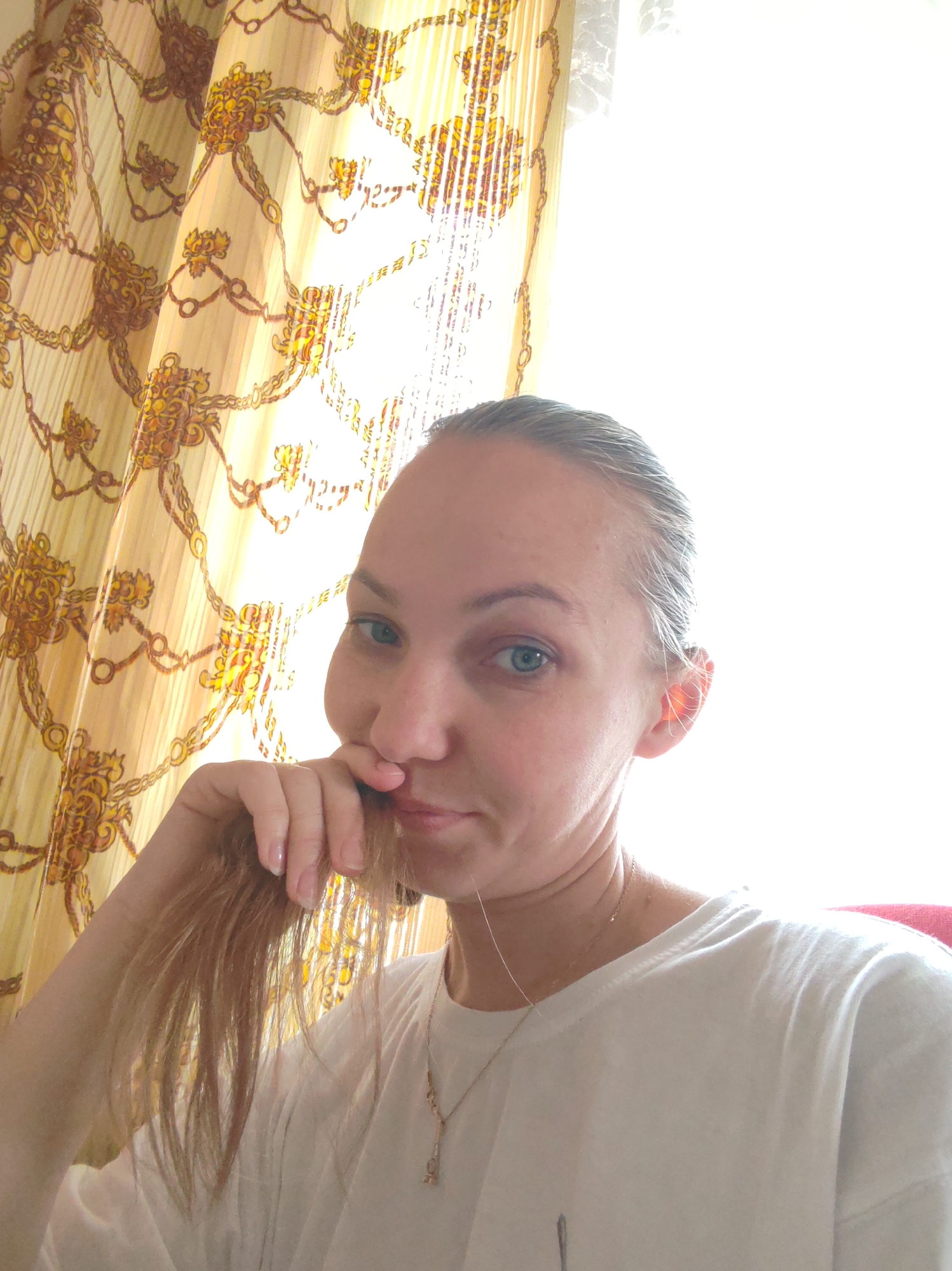 A woman with light hair, blue eyes, and a white t-shirt is pictured indoors, likely in a residence, as suggested by the patterned curtains. She is touching her face with her hand and appears to be looking at the camera. A delicate gold necklace is visible around her neck. The location is mentioned as Чухлэм, Russia, but there is no evidence to support that this image was taken in that city. The background includes a bright window and patterned curtains.