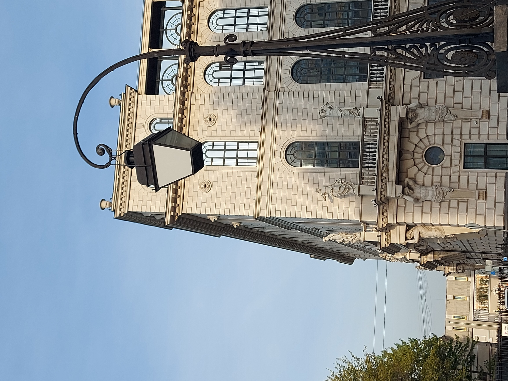 The image shows a street view in Saint Petersburg, Russia, featuring a building facade and a street lamp. The building has a classical architectural style with rectangular and arched windows, decorative moldings, statues, and brick-like facade cladding. A black, ornate street lamp stands out against the sky. The sky is clear. Trees are visible at the bottom of the image. Another building is visible in the distance.