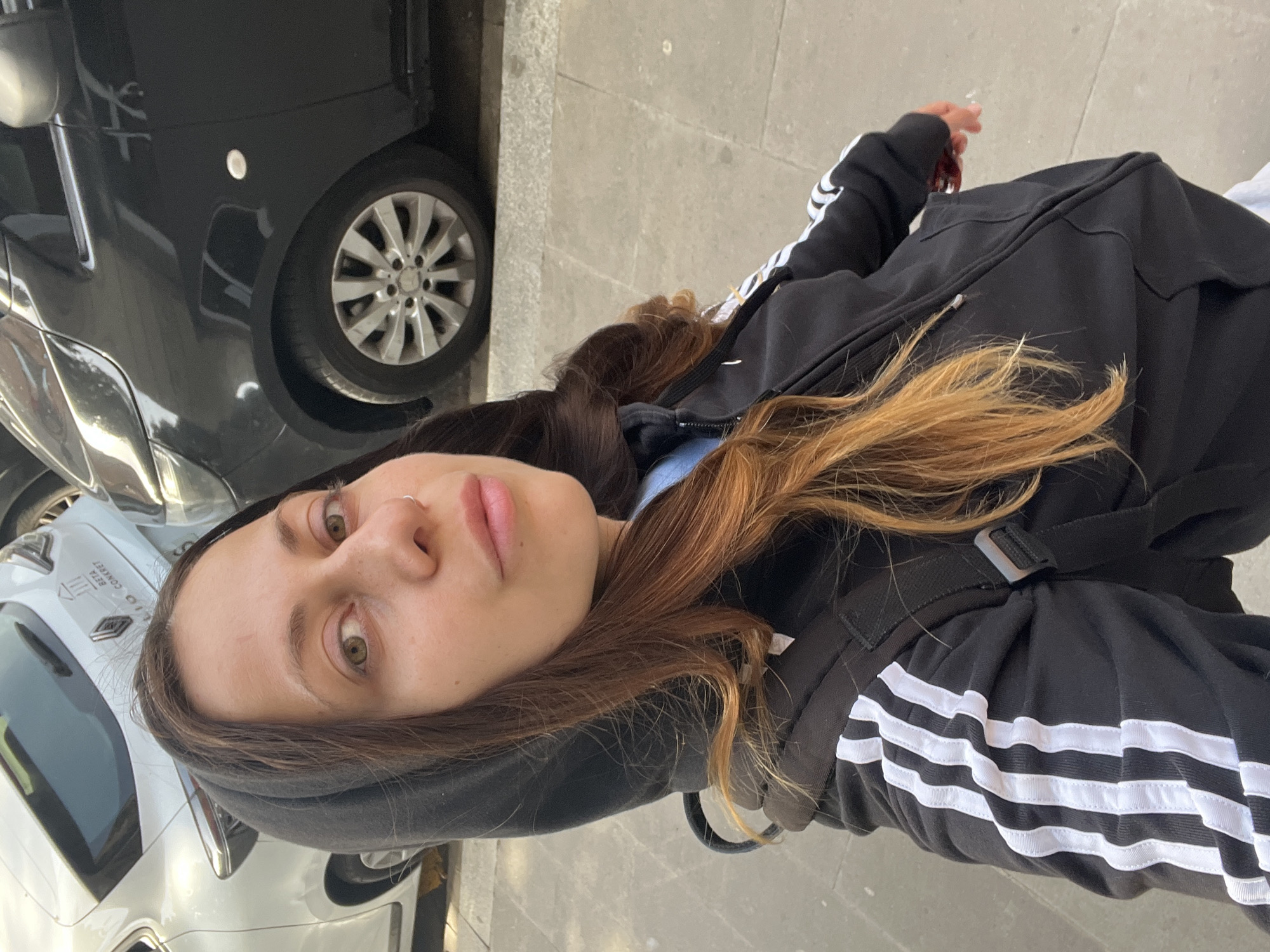 A young woman is depicted in a close-up, chest-up portrait taken on a street in Barcelona, Spain. She is looking directly into the camera with a neutral expression. Her hair is long, dark brown at the roots, and transitions to a lighter, reddish-brown or auburn shade towards the ends. She has green or hazel eyes and a small silver ring piercing her nose.
She is dressed in a black hooded jacket with white stripes visible on the sleeves, characteristic of Adidas apparel. A light blue garment is layered beneath the jacket. Part of a black backpack is visible over her shoulder.
The background features an urban environment with parked vehicles and a grey paved sidewalk. To her left, a dark grey car is visible, identifiable by a wheel with a Mercedes-Benz logo on its hubcap. To her right and slightly behind her, a white Renault car is parked. Text visible on the side of the white car reads "RENT A CAR" and "BETA COMBAT," suggesting it could be a rental or commercial vehicle. The scene is brightly lit by natural daylight, indicating it is daytime.