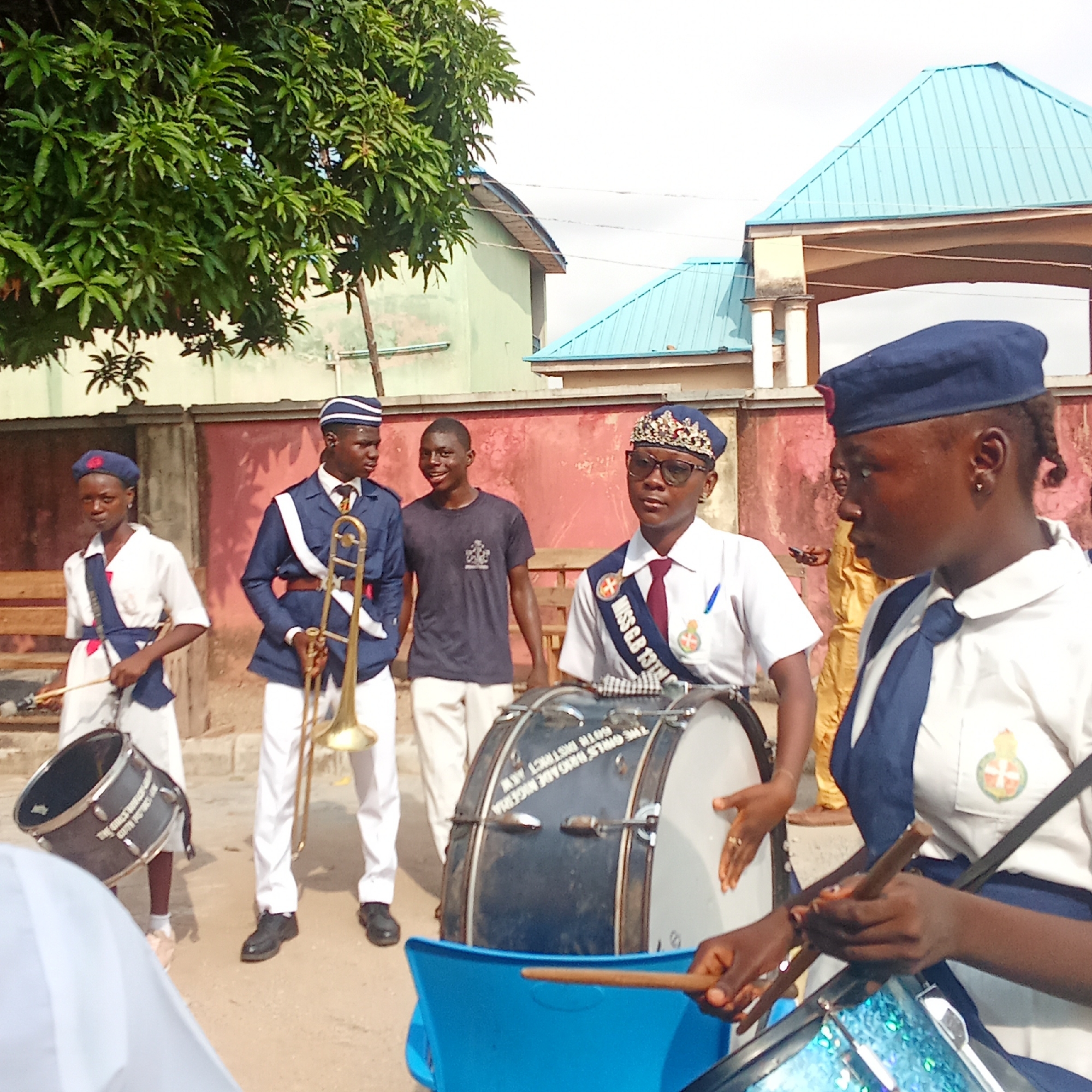In Akwanga, Nigeria, a group of people stand outside, several wearing uniforms indicative of a marching band or youth organization. One person is holding a trombone, while others are positioned with drums, some appearing to be actively playing. There are several buildings visible in the background. The environment is likely an urban or suburban street.