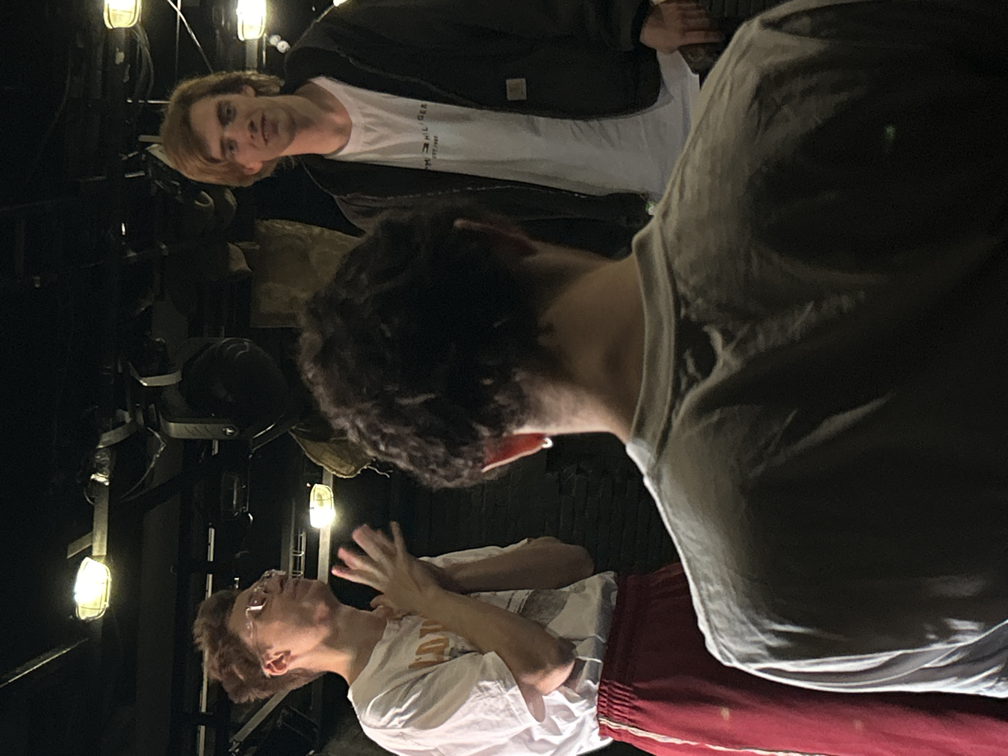 The image shows three young people in a dimly lit setting, possibly indoors or at night. The orientation of the image is rotated 90 degrees counterclockwise.
In the foreground and to the right, a person wearing a gray t-shirt is seen from the back, with their head turned to the left. The person's hair is dark and curly.
To the left of the frame, two people are visible. The person in the middle ground is looking downwards and towards the right. They have light brown hair and are wearing a white t-shirt. Their right hand is raised, with fingers spread, as if in motion.
In the background, another person is seen from the shoulders up, looking towards the left. They have light brown hair and appear to be wearing a white t-shirt under a dark jacket. The text "TOMMY HILFIGER" is visible on the white t-shirt, along with some other smaller text below it.
The background consists of dark shapes and what appear to be industrial lights. Two lights are clearly visible, emitting a warm glow. The overall atmosphere is dark and somewhat mysterious. There are no clear indicators of the specific location or time of day, other than the lighting suggesting it could be evening or indoors. The emotions of the subjects are not explicitly clear, though the raised hand might suggest some form of communication or gesture.