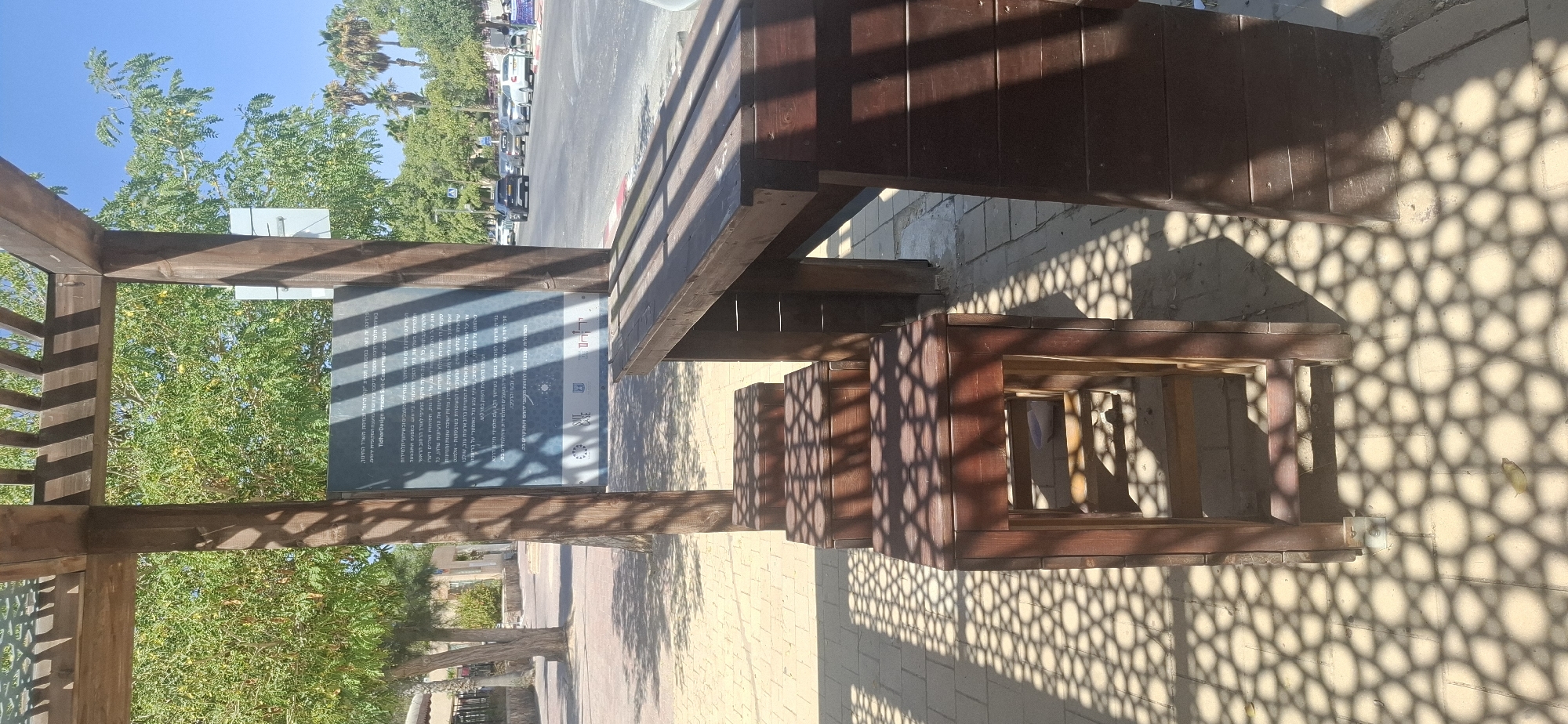 The image captures an outdoor seating area in Yerocham, Israel, beneath a wooden pergola that casts intricate geometric shadows on the tiled ground. Three wooden stools are neatly arranged in front of a matching wooden table, suggesting a place for rest or social gathering. In the background, a sign with Hebrew text, possibly informational or artistic, is partially obscured by a wooden beam from the pergola. Beyond the seating area, a street is visible with parked cars and trees lining the sidewalk, indicating an urban environment. The clear blue sky overhead and the bright sunlight suggest a pleasant day. The overall scene is serene and bathed in the dappled sunlight filtering through the pergola's latticework.