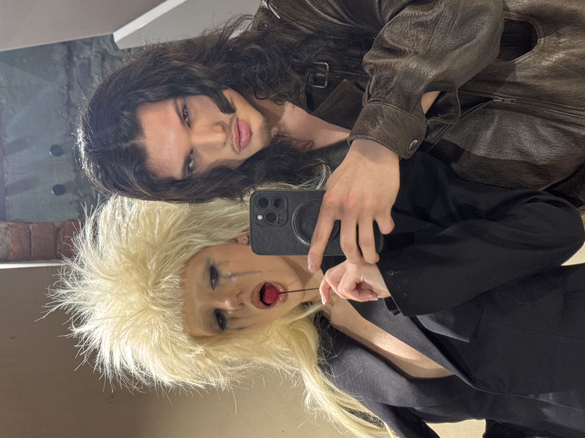 Two individuals are depicted in a close-up, chest-up indoor selfie. The individual on the left, appearing female, has voluminous, spiky blonde hair and dramatic makeup featuring black and silver glitter streaking from under the eyes. She holds a single red raspberry on a thin black stick between her parted lips. She wears a black blazer with a deep V-neck. Her left hand holds a black smartphone, featuring a triple-lens camera system, which is actively capturing the image. The individual on the right, appearing male, has long, dark, wavy hair, pronounced eye makeup, and light facial hair. He wears a dark brown leather jacket with a zippered front and buckle details on the collar. Both individuals are looking directly towards the smartphone's camera lens. The background consists of a plain light-colored wall on the left and a darker, textured wall, possibly concrete or brick, on the right, with a white angular architectural element partially visible on the far right. The specific location within the city of Sofia, Bulgaria, is not discernible from the visual information provided.