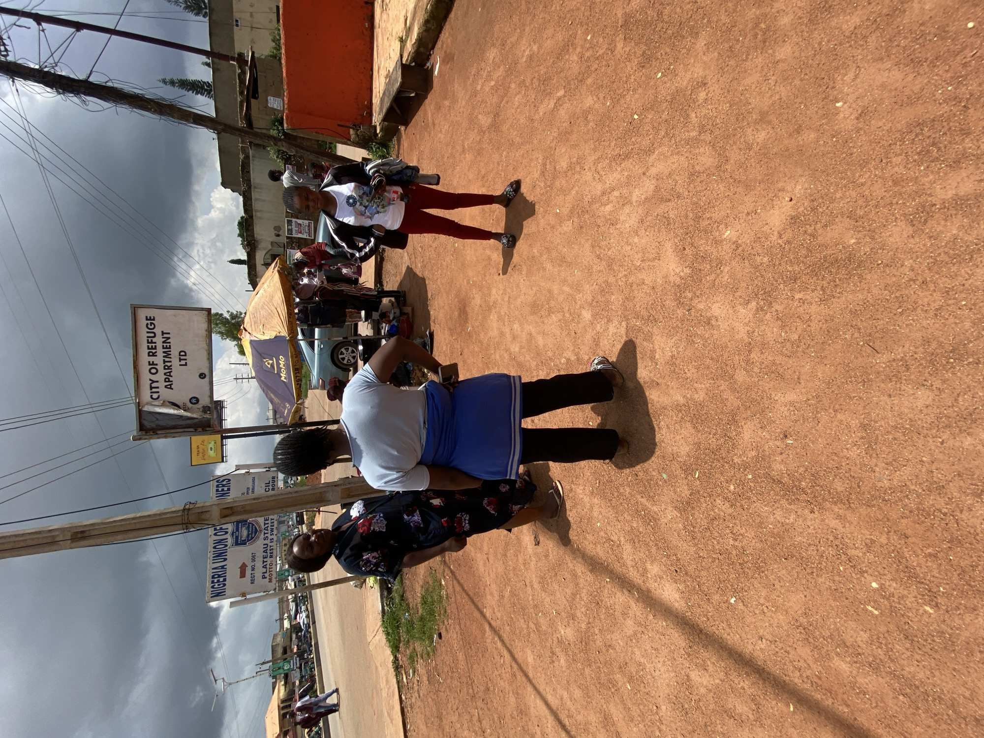 A group of three young women are standing on a dirt road in what appears to be Gura Top, Nigeria. The woman on the left is wearing a dark floral dress and sandals. The woman in the middle is wearing a light blue t-shirt, black pants, and a blue sash tied around her waist. She is facing away from the camera. The woman on the right is wearing a white t-shirt with a graphic print, dark red pants, and sandals. She is holding a phone in her hand. Behind the women, there are signs indicating "Nigeria Union of Pensioners" and "City of Refuge Apartment Ltd." A yellow umbrella is set up, and there are cars and people in the background, suggesting a busy street or market area. The sky is cloudy and overcast.