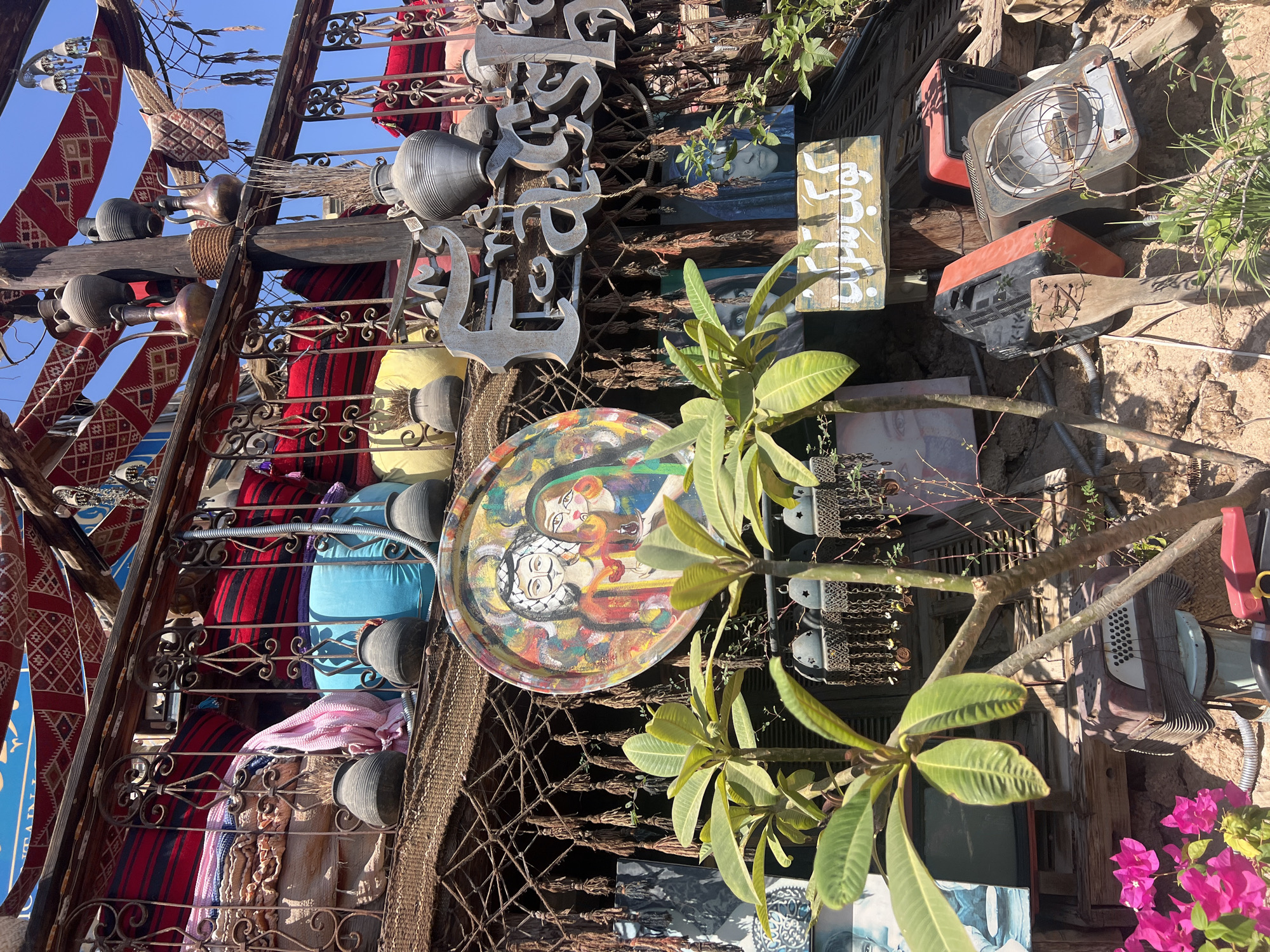 The image captures a vibrant, cluttered display in Sharm El Sheikh, Egypt, likely outside a shop or cafe. The scene is rich with cultural elements and decorative items. In the foreground, lush green plants with broad leaves dominate, with bright pink flowers visible in the lower left corner. Behind the plants, various objects are artfully arranged.
A circular, painted tray featuring a portrait of a couple with distinct facial features and traditional attire hangs prominently. Above this, metal lettering spelling out "Fashinista" is affixed to a wooden beam. Further up, traditional Arabic textiles with red and geometric patterns are draped, providing a colorful canopy.
Several clay pots and jugs of various sizes and designs are displayed on wooden shelves and railings. Metal lanterns with intricate designs hang from chains. In the background, portraits of faces, one with striking blue eyes, are visible, adding to the artistic and eclectic atmosphere.
At the bottom of the image, several discarded or repurposed items are visible, including an old television set, a space heater with a wire grill, and what appears to be an accordion. A wooden sign with Arabic script is also present. The overall impression is a charming, bohemian-style outdoor space decorated with a mix of traditional and contemporary elements, indicative of a marketplace or artisan shop in Egypt.