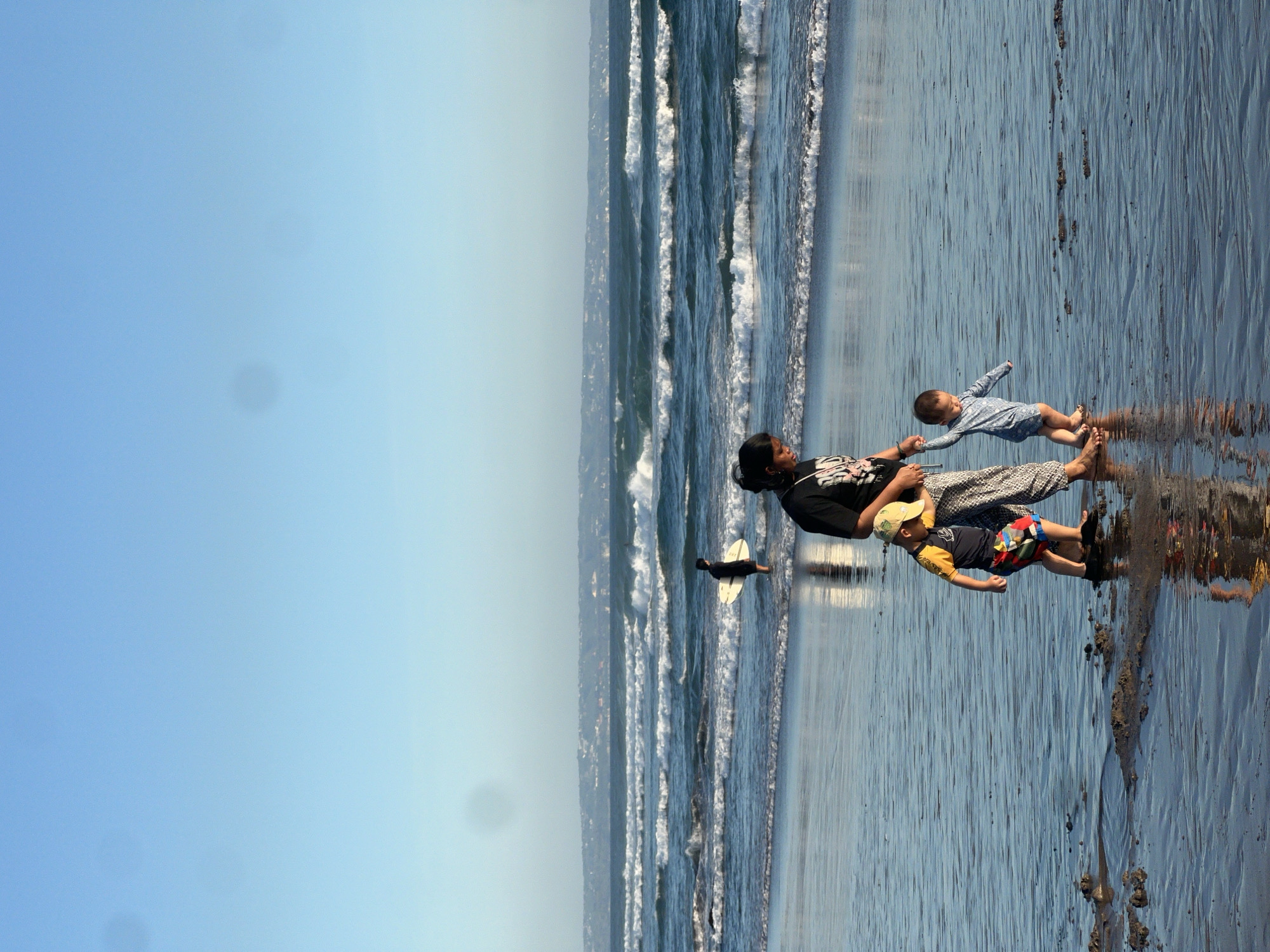 In North Kuta, Indonesia, on a beach with calm, shallow waves lapping the shore, a woman wearing a black t-shirt and patterned pants walks with two young children. To the left, a boy in a blue and yellow rash guard and colorful shorts, wearing a yellow cap, holds the woman's hand. To the right, a baby in a blue long-sleeved romper also holds the woman's hand, appearing to take a step. In the background, a person in a black shirt and shorts stands in the water holding a white surfboard. The sky is clear blue with a hint of haze on the horizon, where buildings are faintly visible. The wet sand reflects the sky and the figures on the beach.