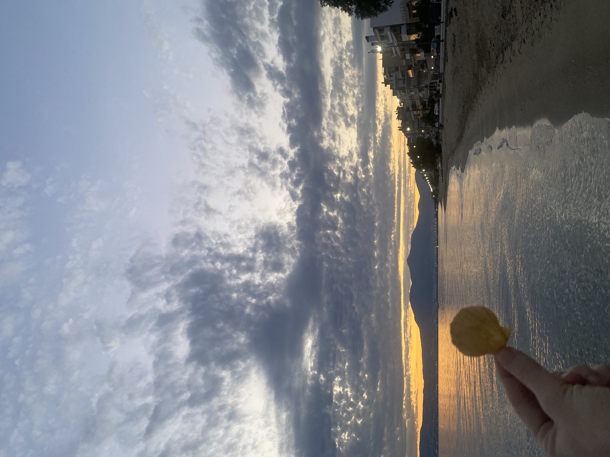 A hand holds up a small, round, golden-brown object, possibly a piece of food, against a seascape at dusk. The object is in focus, while the background is slightly blurred. The sea reflects the colors of the sky, with shades of orange and yellow from the setting sun on the horizon, and blues and grays from the clouds. In the distance, a silhouetted mountain range stretches across the horizon. Along the coastline, a beach is visible, with sand leading up to a row of multi-story buildings. Streetlights are illuminated along a road that runs parallel to the beach and buildings. The overall impression is a serene, end-of-day scene in a coastal town. The location is Neoi Epivates, Greece.