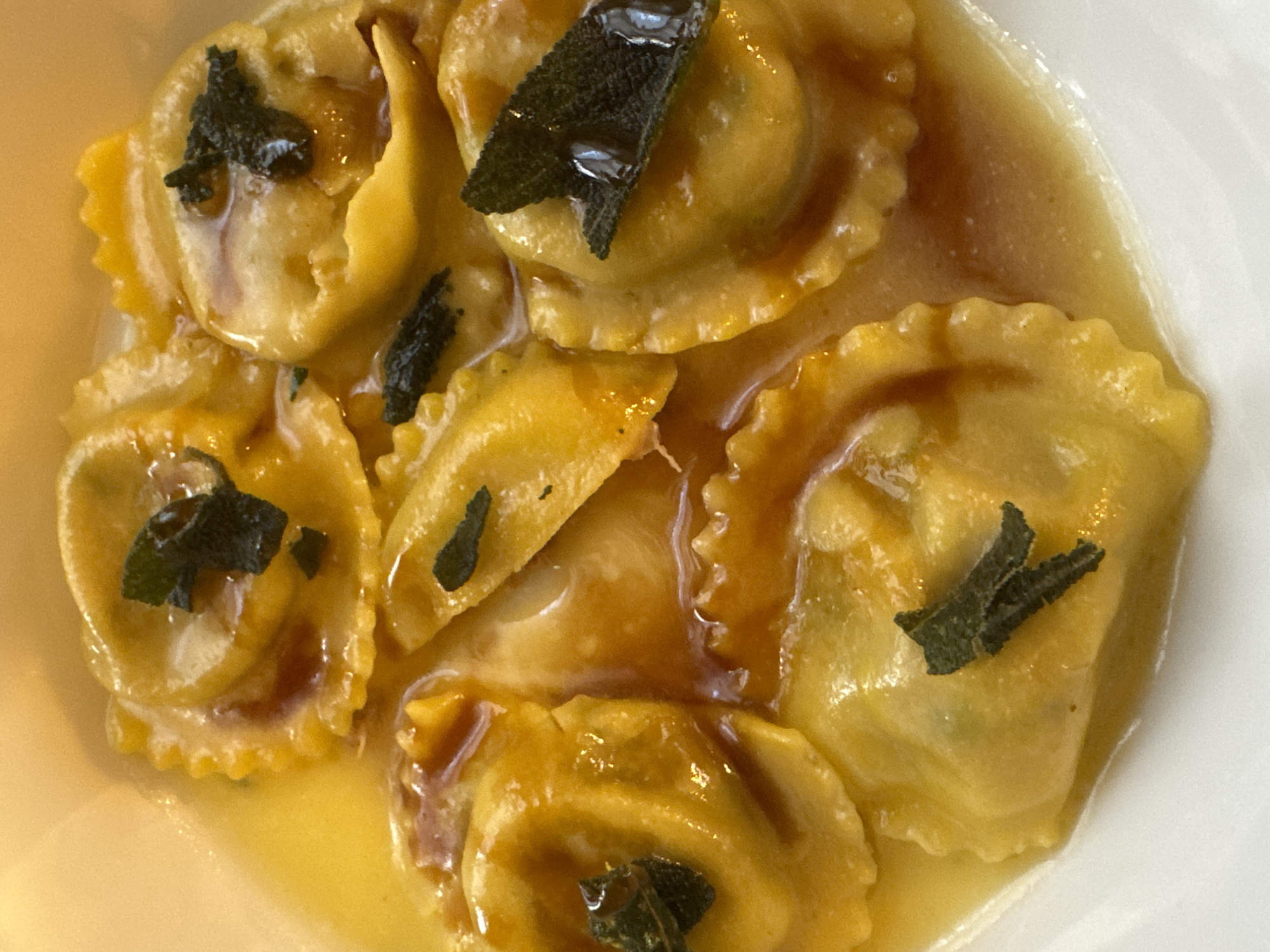 The image presents a close-up, overhead view of a serving of pasta in a white, shallow bowl or plate. The dish consists of approximately eight to nine pieces of yellow-orange, crimp-edged filled pasta, resembling ravioli or tortelli. These pasta pieces are bathed in a glossy, golden-brown, rich-looking sauce. Several individual dark green sage leaves, appearing slightly oily, are placed on top of various pasta pieces. A subtle green hue is visible beneath the pasta dough in some areas, indicative of a filling. No individuals, actions, or interactive elements are depicted. The specific location within the city of Milan, Italy, cannot be determined from the visual information provided in the image.