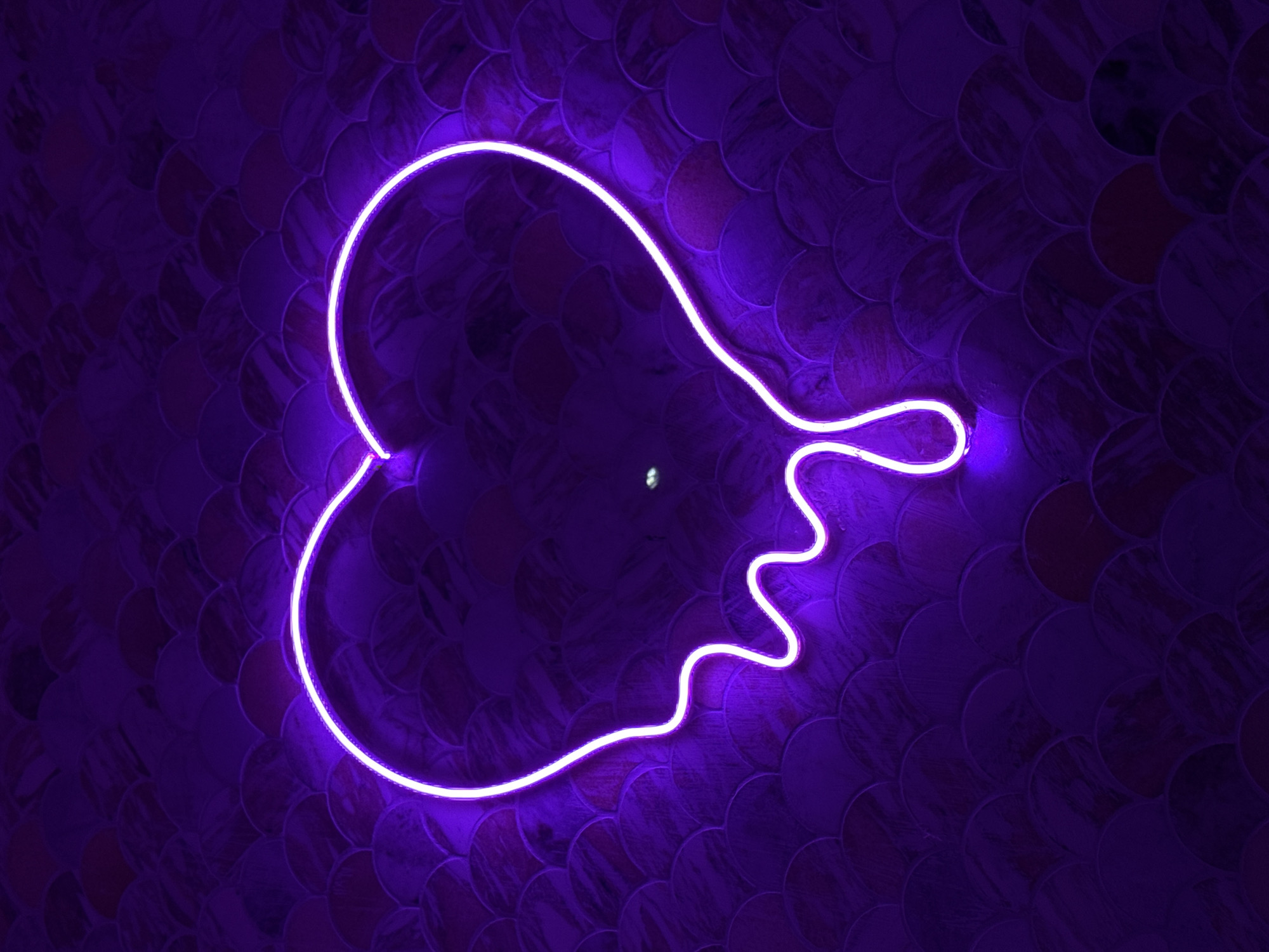 A neon sign, glowing a vibrant purple, dominates the scene. The sign is in the shape of a stylized face or head with flowing hair or tendrils. The sign is mounted on a textured wall. The wall appears to be tiled with small, overlapping circular shapes that are shades of dark purple. There is a single, bright reflection near the center of the image. The color palette is limited to shades of purple, creating a monochromatic and atmospheric effect. The image suggests an interior location, potentially a bar, club, or art installation in Tel-Aviv, Israel.