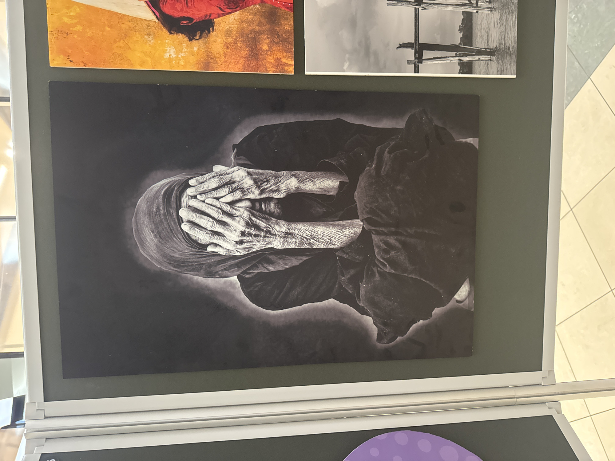 The image displays an exhibition of photographs mounted on a dark green wall. The main photograph, in black and white, features a close-up of an elderly person's hands covering their face, with a headscarf visible. To the right of this image, another photograph shows a person wearing red against an orange background, and below that, a black and white photograph depicts a wooden pier extending into water with a figure standing on it. The exhibition is set in a well-lit indoor space with a tiled floor. The overall scene suggests an art exhibition. The location of this exhibition in Kilkenny, Ireland is not discernible from the visual information provided.