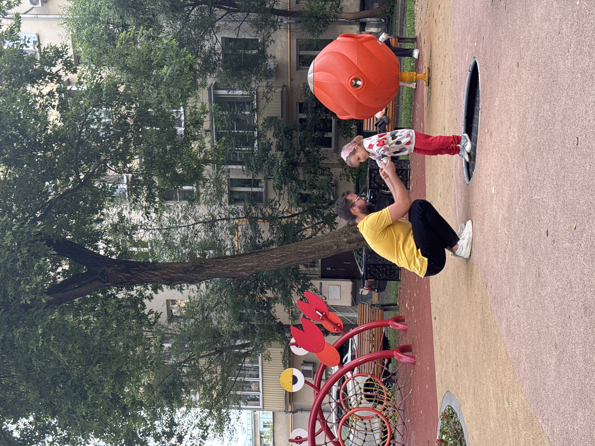 A father and his young daughter are on a playground in Moscow, Russia. The father, wearing a yellow t-shirt and black pants, is crouched down and holding his daughter's hands. The daughter, wearing a white sweater with hearts and red pants, is standing on a trampoline. The playground has red and tan colored soft surfaces. In the background, there is a large tree, a modern apartment building with balconies, and a red playground structure with a net climber and red abstract shapes. There is also a large, orange, egg-shaped play structure with a grey top and another person sitting on a bench in the background. The scene appears to be a typical urban playground in Moscow.