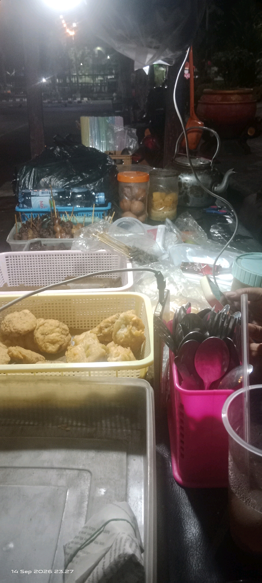 The image depicts a street food stall at night in Klaten, Indonesia. In the foreground, a yellow plastic basket contains fried food items, possibly chicken or fish. To the right, a pink plastic container holds an assortment of black plastic cutlery, including spoons and forks. Next to it, a clear plastic cup with a dark liquid and ice is visible. A large, translucent plastic container with a newspaper wrapped around an object is positioned in the lower left corner. In the background, various items are arranged on a table, including a kettle, jars containing food, and packages of food on skewers. A black garbage bag is also present. The scene is lit by a bright overhead light, casting shadows and creating a hazy atmosphere. The background suggests an outdoor setting with trees and buildings faintly visible.
