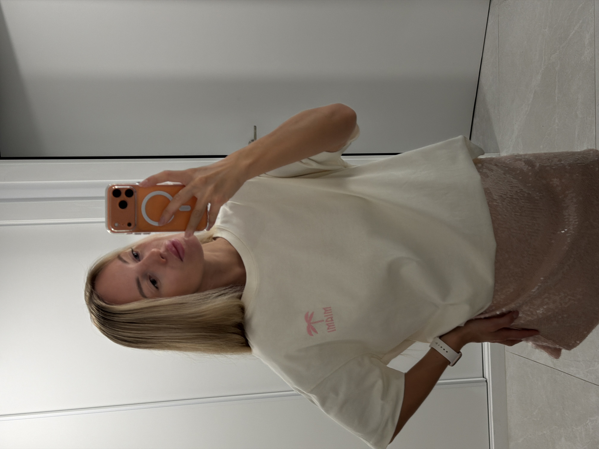 The image shows a young woman taking a selfie in what appears to be a modern, well-lit indoor space. She has blonde hair and is wearing a cream-colored t-shirt with a small pink graphic of a palm tree and the text "MIAMI" below it. Her skirt is shimmering and appears to be made of sequins, in a rose gold or light pink color. She is also wearing a white smartwatch on her left wrist.
The woman is holding an orange iPhone with a clear case, angled to capture her reflection in a mirror or reflective surface. Her expression is neutral, and she is casually posing with her left hand on her hip. The background consists of smooth, light-colored walls, possibly marble tiles on the right, and a white door or closet on the left. The lighting suggests it is daytime, and the overall atmosphere is clean and contemporary. There are no explicit cues about the weather or time of day, but the indoor setting implies it is not dependent on external conditions. The presence of the phone and the selfie pose indicate she is documenting herself. The overall impression is of a person showcasing their outfit in a stylish, private setting.