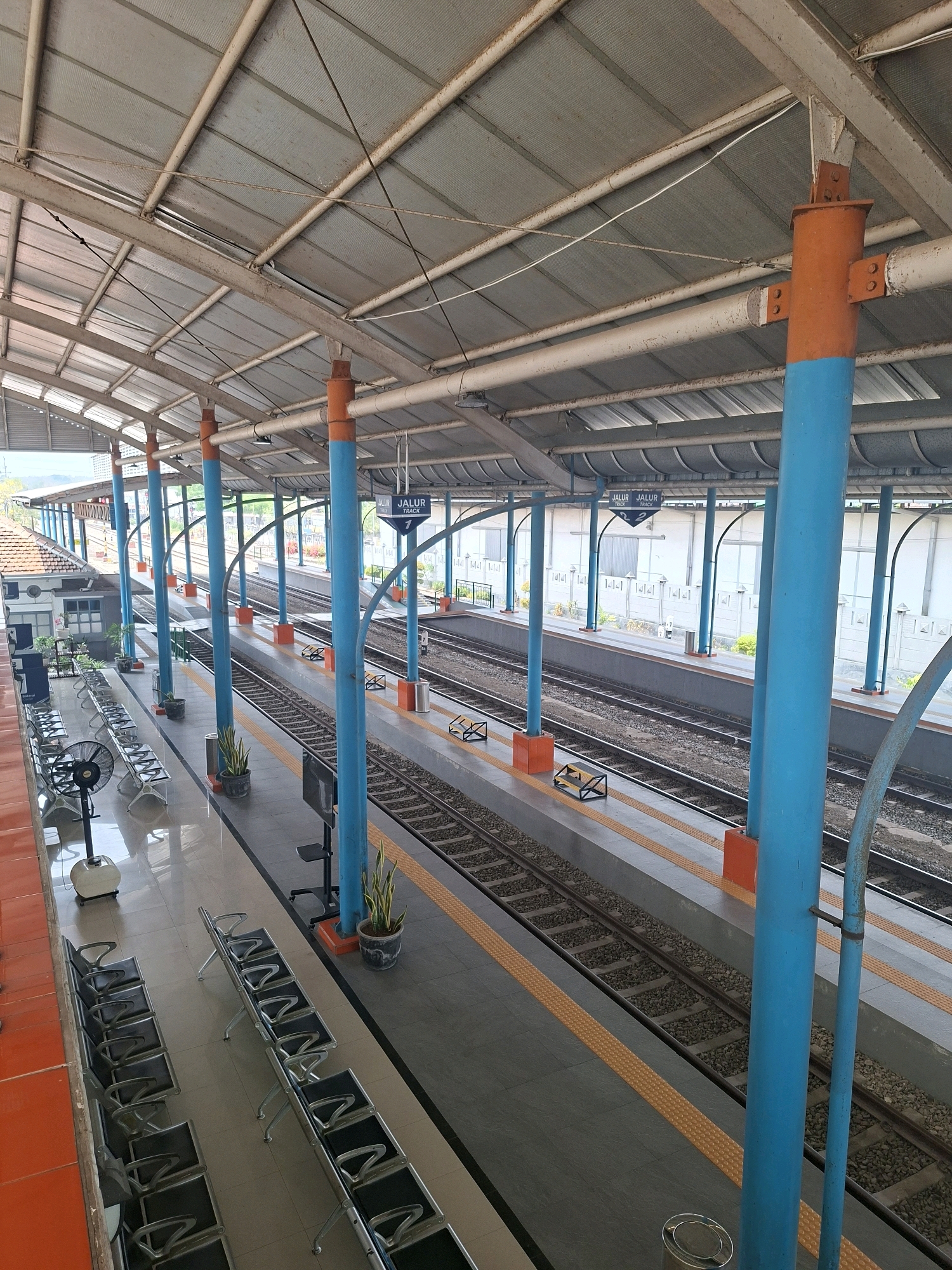 This is a photograph taken at a train station in Depok, Indonesia, likely Depok Old Town Station (Stasiun Depok Lama) or Depok Station (Stasiun Depok). The image captures a view of the train tracks and platforms from an elevated perspective.
The scene is dominated by a covered walkway supported by numerous columns painted in a vibrant blue and orange. The roof of the walkway is made of corrugated metal sheeting. Train tracks run horizontally across the frame, with platforms on either side. The platforms are paved with grey tiles and have orange tactile paving near the track edges.
Several rows of empty metal benches are arranged on the platforms, indicating a waiting area for passengers. A pedestal fan is visible on the left side of the platform, positioned near some of the benches. Potted plants are placed sporadically on the platforms.
Signage indicating "JALUR TRACK" (Track Line) followed by numbers such as "1" and "2" are suspended above the platforms, helping passengers identify the correct track. In the background, there are white buildings with some architectural details, and glimpses of greenery suggest trees and vegetation outside the station area. The overall impression is that of a functional, open-air train station environment. There are no people visible in the photograph.