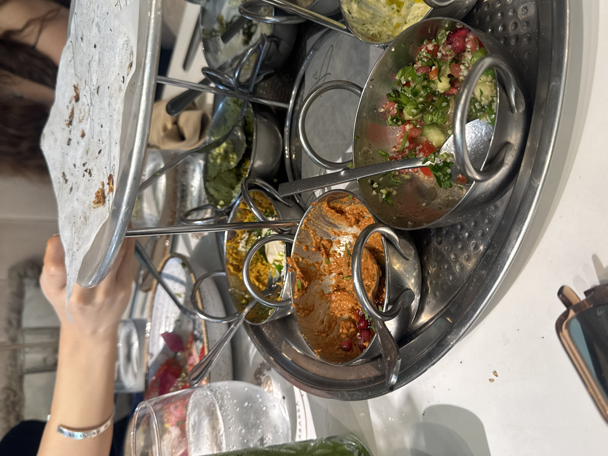 In Aventura, Florida, a close-up shot captures a metal tray laden with small bowls of assorted dips and salads. The tray sits on a white table, with a glass of water and a green beverage bottle visible in the background. A person's arm, adorned with a silver bracelet, is seen reaching towards the tray, suggesting active dining. The dips appear to be various shades of orange and green, with some containing visible ingredients like pomegranate seeds and chopped herbs. The overall setting suggests a casual dining experience, possibly at a restaurant.