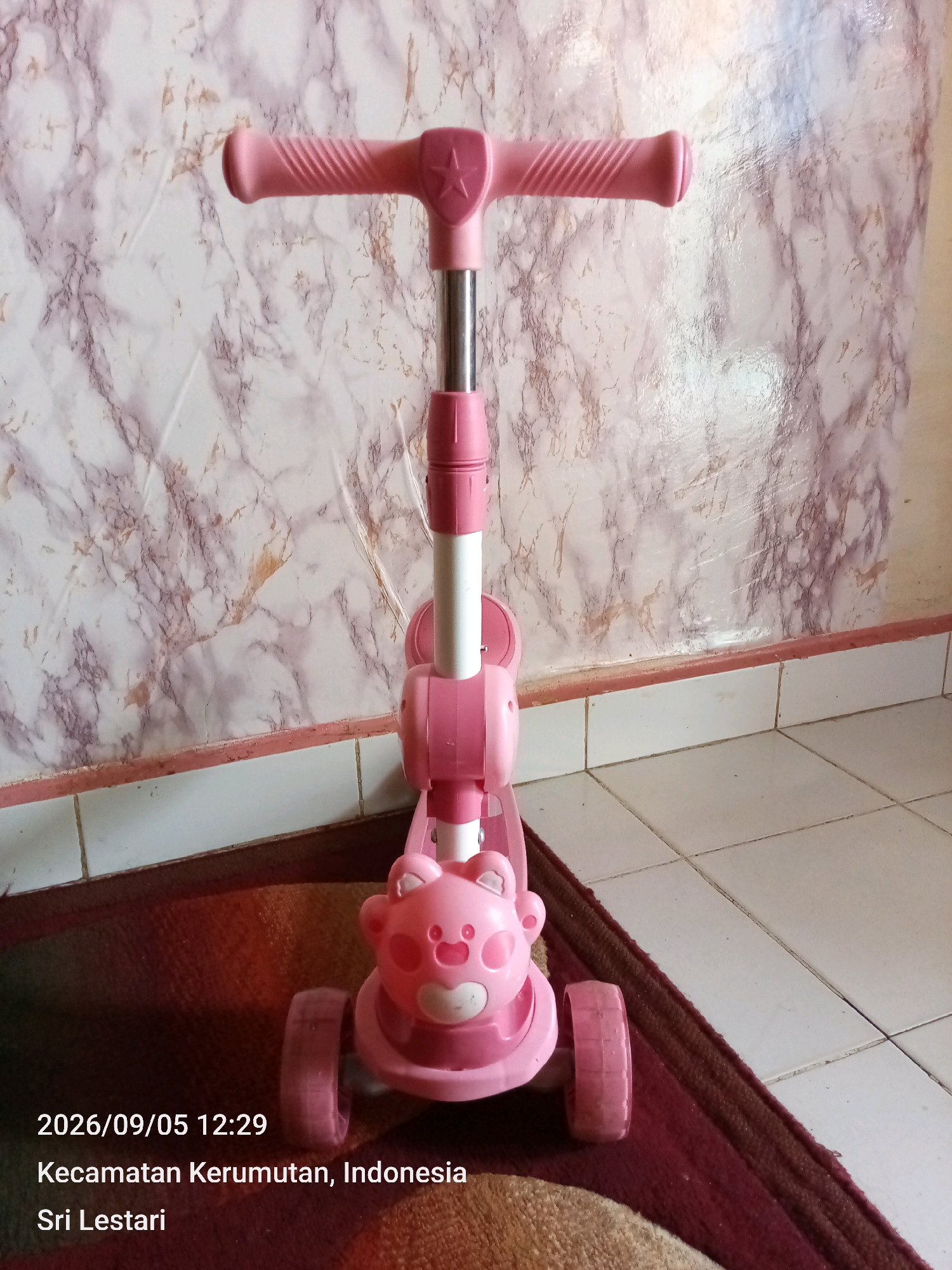 A pink children's scooter is positioned on a rug in front of a marble-patterned wall. The scooter has a pink handlebar with grips and a star emblem, a white and pink adjustable stem, and a pink base shaped like a bear's face with pink ears. It has two pink wheels visible at the front and a third pink wheel at the rear. The floor tiles in the background are cream-colored. Text superimposed on the lower left of the image indicates the date and time as "2026/09/05 12:29" and the location as "Kecamatan Kerumutan, Indonesia Sri Lestari."
