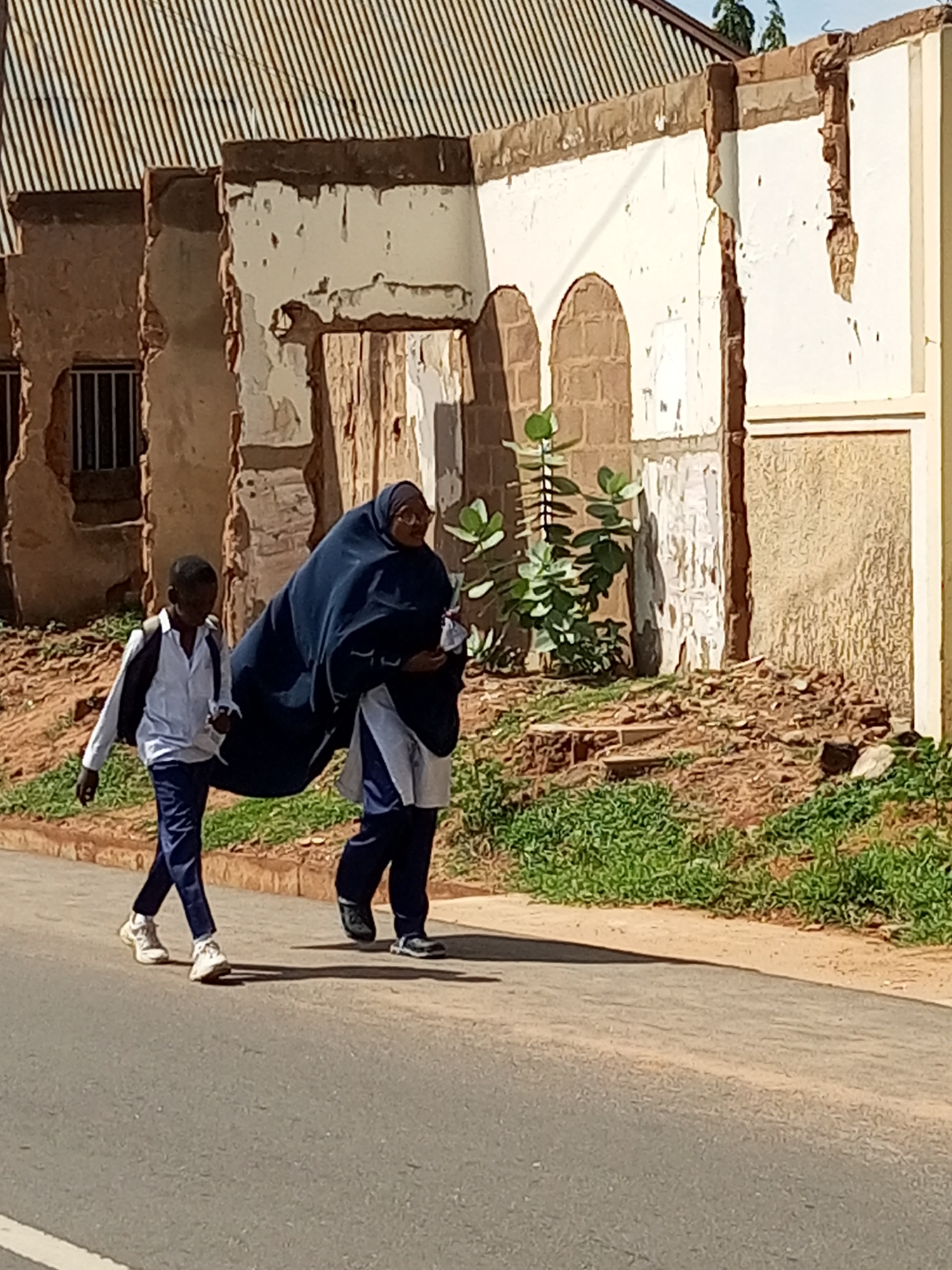 Two individuals are walking on a paved road in front of a dilapidated building in Gombe, Nigeria. On the left, a young boy wearing a white shirt, dark pants, and a black backpack walks forward. To his right, a woman is partially obscured by a large, dark blue cloth draped over her head and shoulders. She is wearing glasses and a white garment underneath the cloth. The building behind them appears to be under construction or renovation, with exposed brickwork and peeling paint. Green vegetation grows along the side of the road and around the base of the building. The scene is captured in daylight under a clear sky.