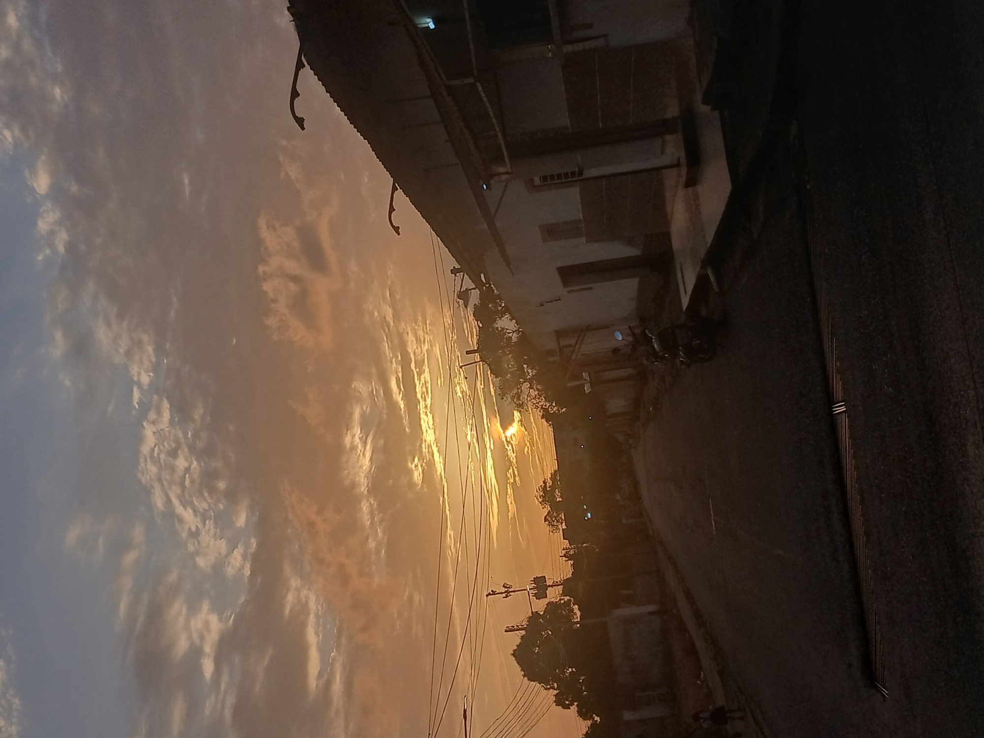 The image presents a street scene at sunset in what could be Juan Griego, Venezuela, though specific landmarks are not visible to confirm the location. A building with a corrugated roof and several windows lines the left side of the frame. On the right is a paved road, partially obscured by deep shadows. A motorcycle is parked near the building. Power lines stretch across the sky, silhouetted against the golden sunset and scattered clouds. The sun's light casts a warm glow over the scene, illuminating trees and smaller buildings further down the road. The overall atmosphere suggests dusk or early evening.