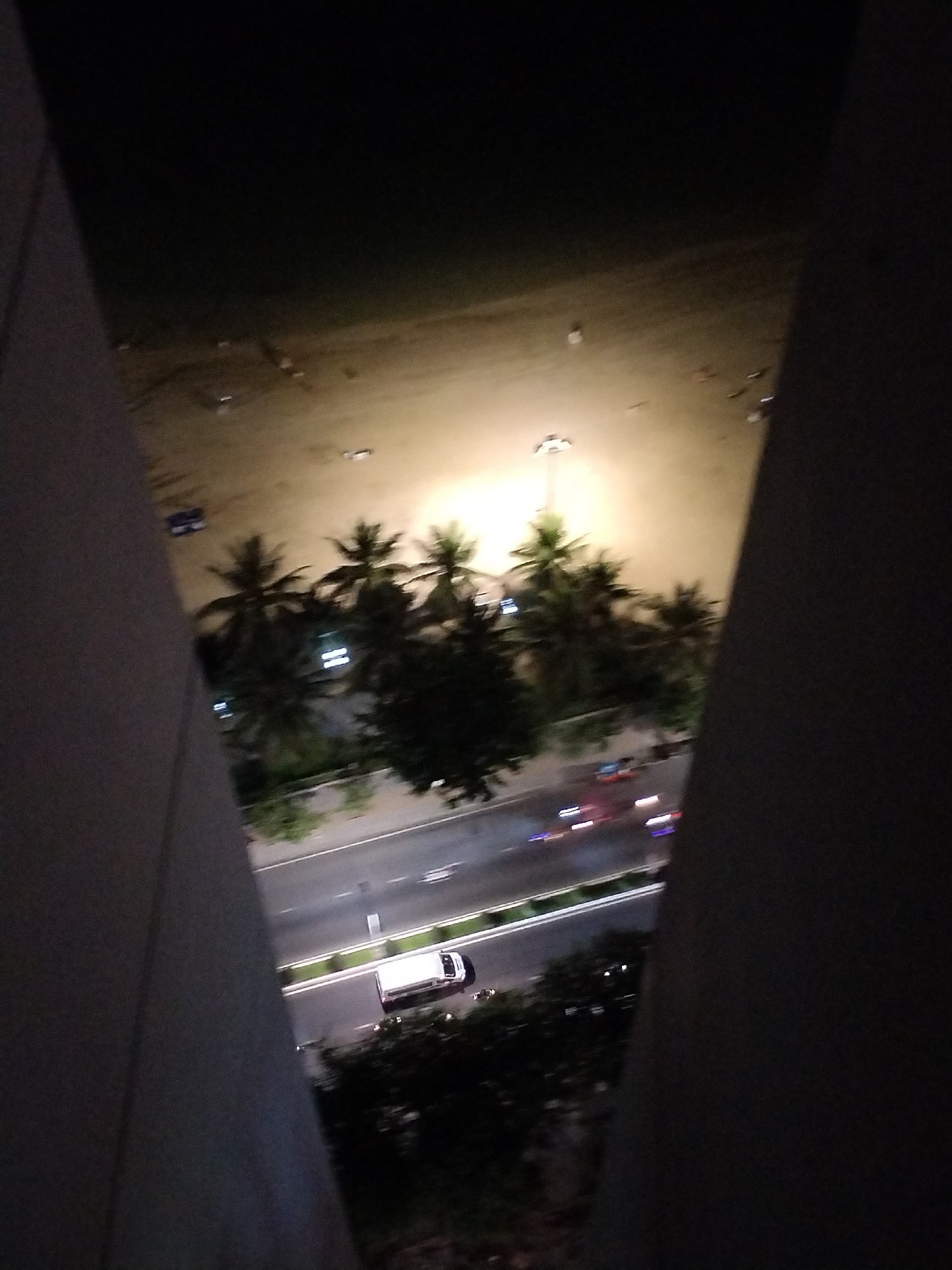 The image, taken at night, depicts a view from a high vantage point, likely a building in the Bac Nha Trang Ward of Vietnam. The composition is framed by the building's structure, creating a triangular opening. The scene includes a beach illuminated by a light source, palm trees, a road with moving vehicles represented by light trails, and a white van. The overall environment is dark.