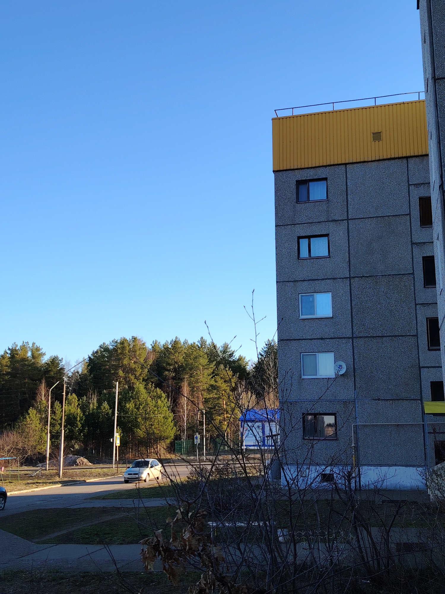 This image captures a clear day in Markovskii, Russia, showcasing a residential building, a road, and a dense forest under a bright blue sky.
The most prominent feature is a multi-story concrete panel building, characteristic of post-Soviet residential architecture. Its facade is composed of large, gray concrete slabs, and the top section is strikingly covered in bright yellow corrugated metal siding, topped with a dark railing. Various windows are visible along its height, some reflecting the sky, while a satellite dish is mounted near a lower-level window. The lower part of the building's foundation has been painted light blue and white.
To the left and across the background, a dense forest of evergreen pine trees provides a natural backdrop, suggesting the proximity of the residential area to a wooded landscape. Several streetlights or utility poles are visible along the edge of this forest, near the road.
In the middle ground, a paved road or driveway curves through the scene. A light-colored sedan is either parked or driving on this road, partially illuminated by the sun. A yellow road sign is visible in the distance near the vehicle. A small blue and white structure, possibly a utility building or kiosk, can be seen closer to the base of the main building, partially obscured by foreground branches.
The immediate foreground is dominated by the bare, intricate branches of deciduous trees and shrubs, indicative of late autumn or early spring. Patches of dry grass and fallen leaves cover the ground.
The sky is a brilliant, cloudless blue, filling the upper portion of the frame. The lighting suggests either early morning or late afternoon, with the sun casting gentle illumination on parts of the scene. The overall atmosphere is quiet and still. There are no people visible, and no discernible text can be read on any objects.