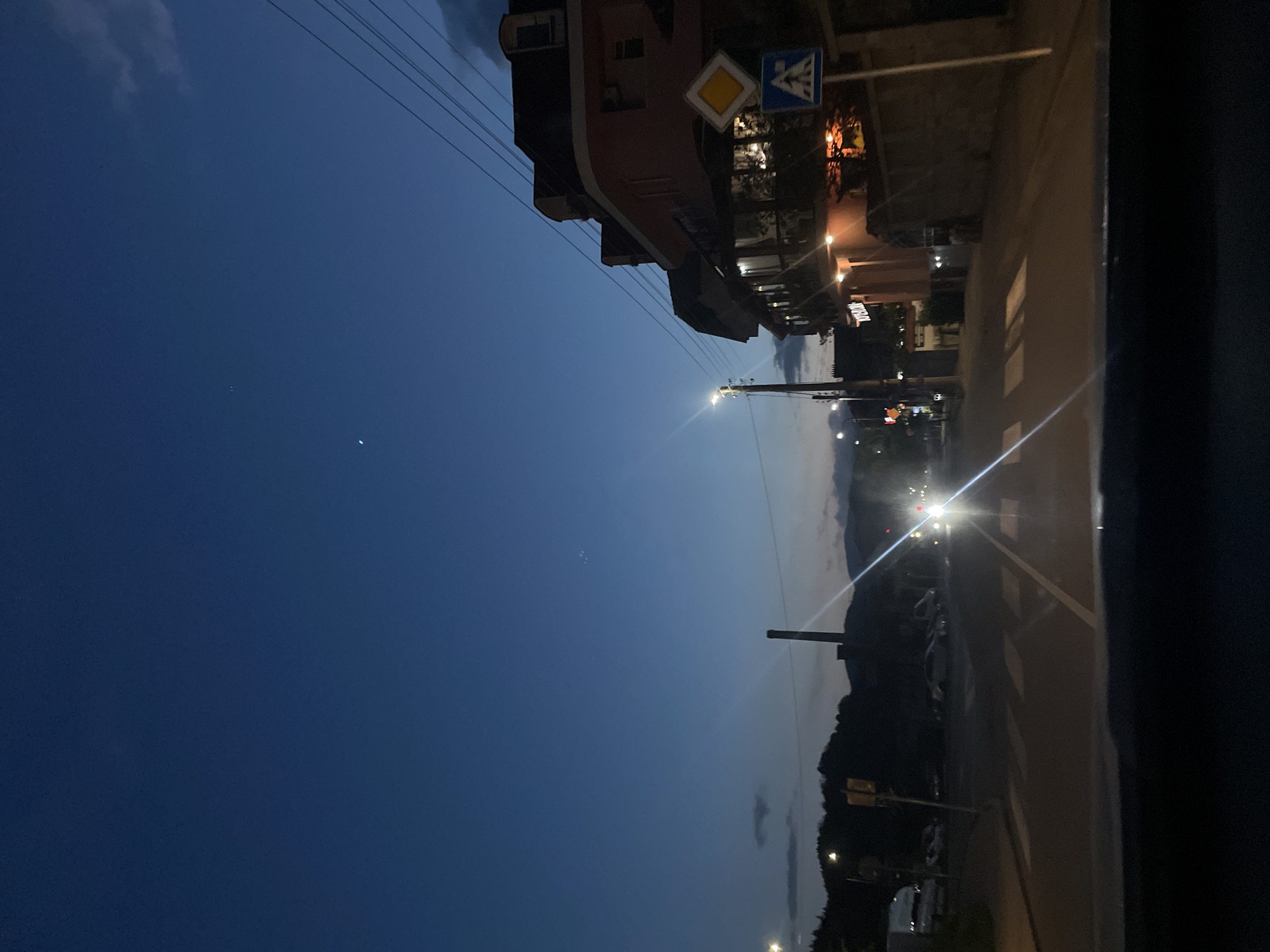 The image shows a night scene in the city of Zlatograd, Bulgaria. The photograph is taken from a high angle, possibly from a window or elevated position. The sky is a gradient of deep blue, transitioning into a lighter blue-gray towards the horizon. Power lines are visible crossing the upper portion of the image.
On the right side of the image is a road with white markings, suggesting a crosswalk. A building with illuminated windows stands along the road. Signage, including a pedestrian crossing sign and a yellow diamond-shaped sign, is mounted on poles near the building.
Streetlights illuminate the road and surrounding area. In the distance, a darker silhouette of a mountain range is visible. The overall impression is that of a quiet evening in a small town.