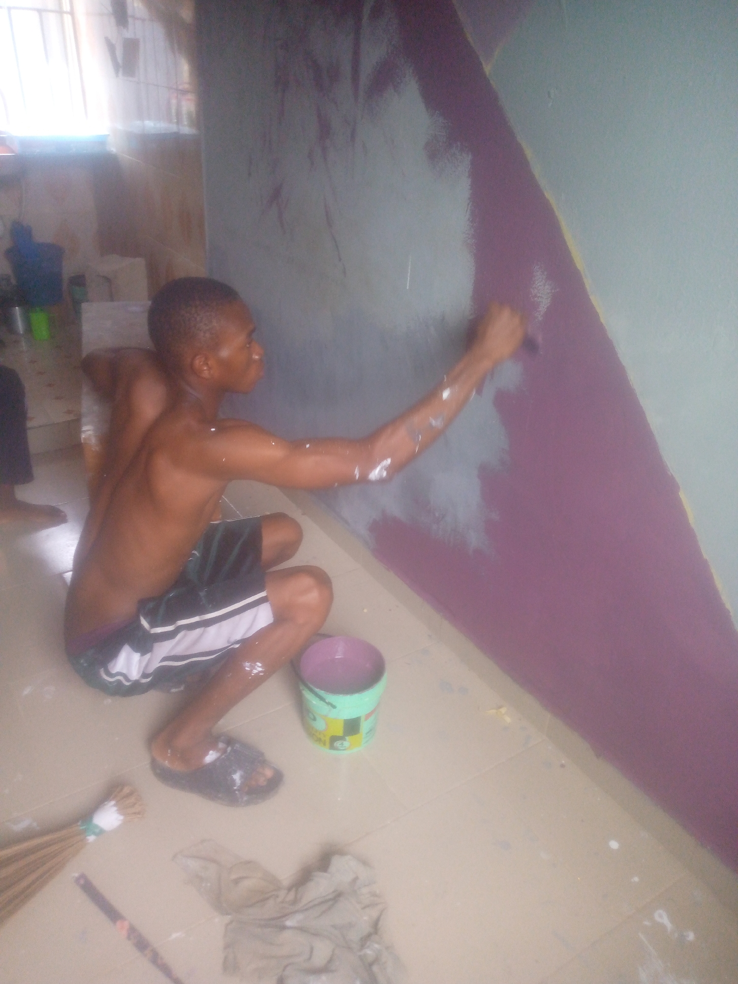 The image shows a young man actively painting an interior wall while crouching on a tiled floor. He is shirtless, wearing dark shorts with white stripes, and dark sandals. His right arm and shoulder are visibly splattered with paint. He holds a paintbrush in his right hand, applying a light grey or white paint onto a section of the wall that is partially painted purple or dark pink. A distinct diagonal yellow line separates the two colors on the wall, possibly indicating a design boundary or masking tape.
Next to the painter, a light green bucket containing purple paint is on the floor. Faint text "QUALITY" and the number "4" are discernible on the side of the bucket. The floor, composed of light beige tiles, is dotted with various paint splatters. In the foreground, near the man's left foot, a natural fiber broom tied with a white and green band lies on the floor, alongside a paint-stained light brown cloth or rag.
In the blurred background to the left, the leg and foot of another person are partially visible. Further back, there are indistinct objects, including what appears to be a blue bucket and a white stool. Bright light streams in from a window with vertical bars, illuminating the scene and suggesting it is daytime. The man appears focused on his task.