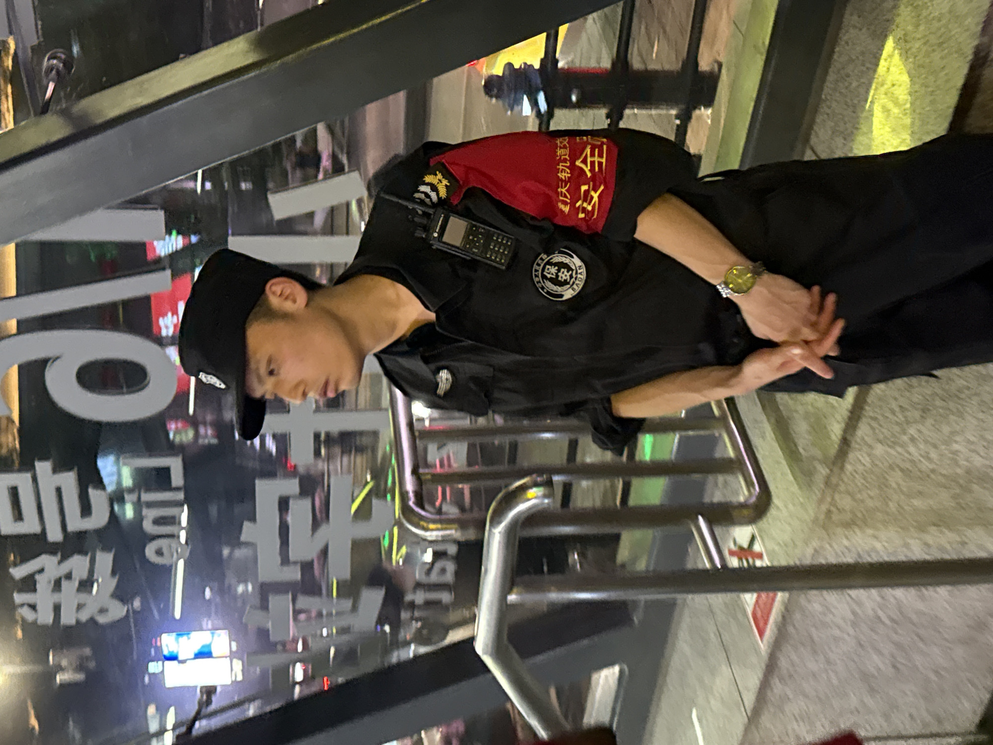 A security guard stands outside in Yuzhong District, China. The man wears a black uniform with a red armband labeled “Chongqing Rail Transit Safety” in Chinese. He also wears a black cap and a watch on his left wrist, and holds a radio attached to his chest. The environment includes a reflective glass surface with the word "Club" visible, and a metal railing. The ground is paved.