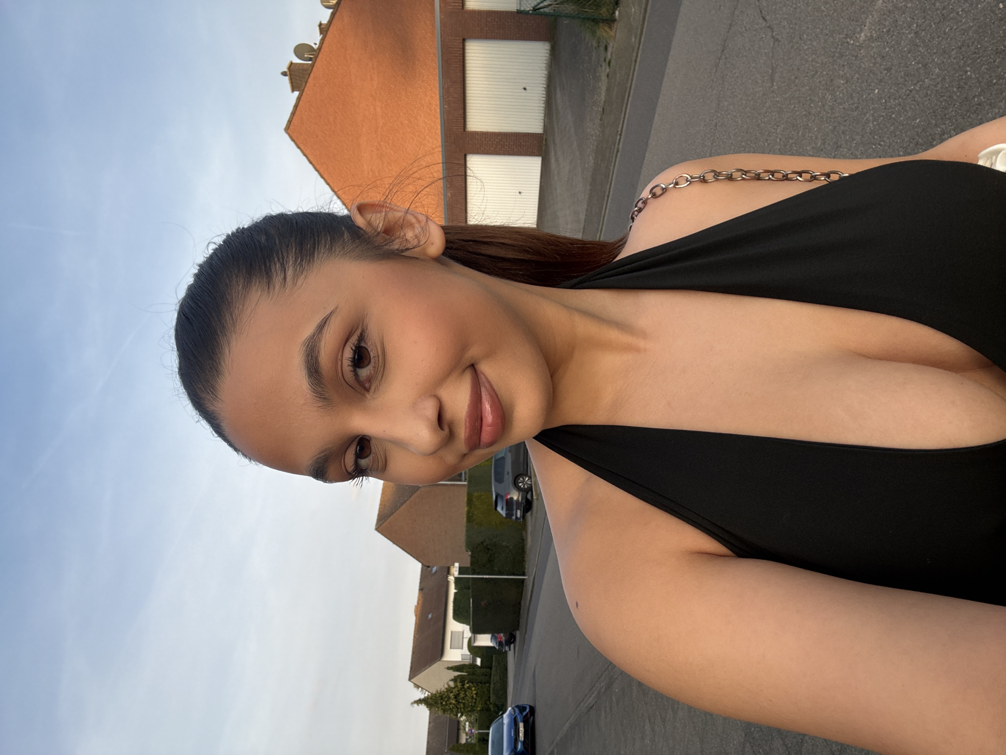 A young woman is taking a selfie outdoors. She has dark hair pulled back from her face, light brown eyes, and is wearing a black dress with a chain strap. The background shows a residential street. There are houses with orange roofs and light-colored garage doors. Cars are parked along the street, and there are trees and bushes lining the sidewalks. The sky is a light blue color. The location is consistent with a residential area within the city of Mönchengladbach, Germany.