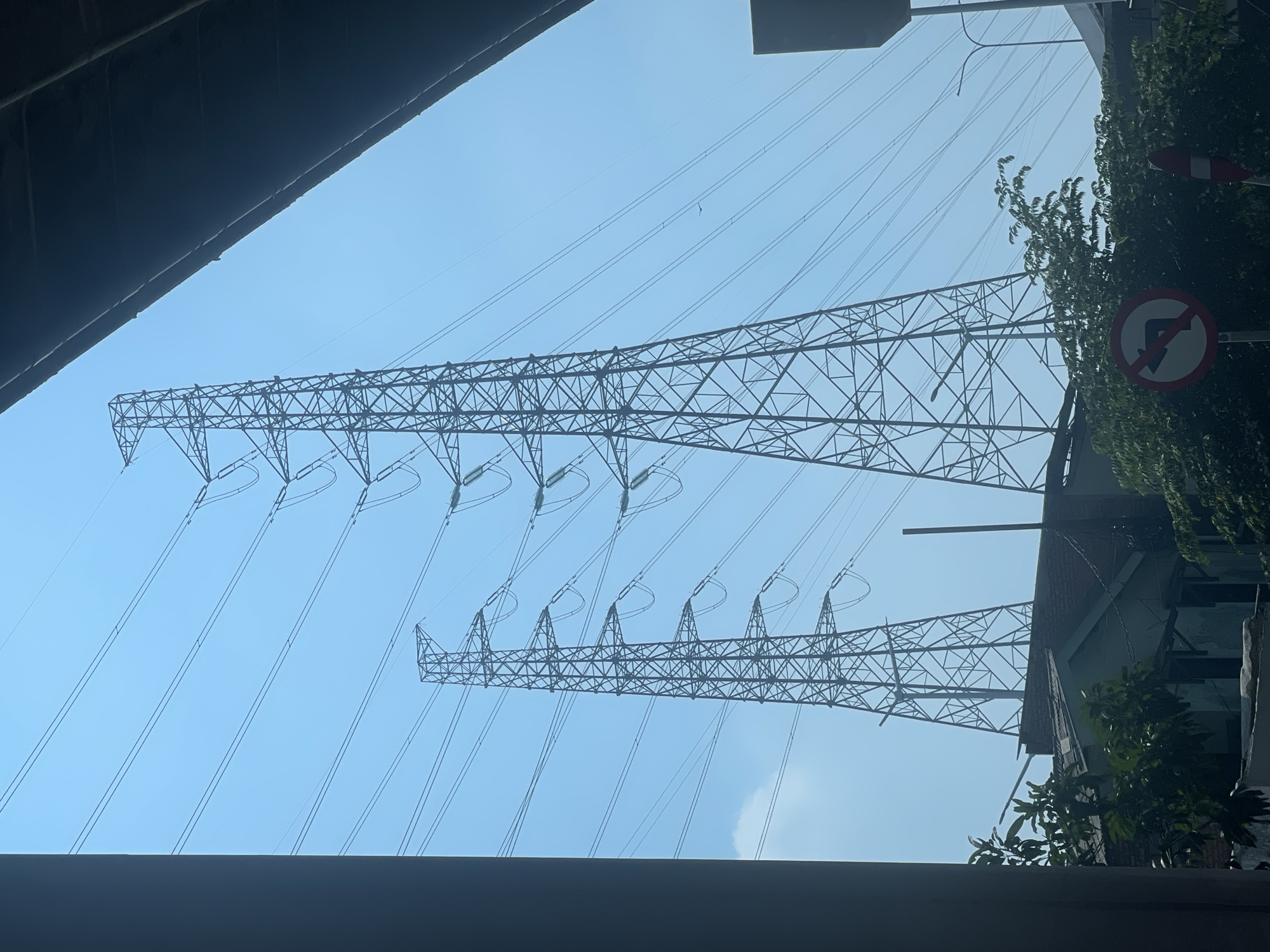 The image captures a segment of the sky in North Jakarta, Indonesia, dominated by two large electricity pylons and a web of high-voltage power lines stretching across the frame. The pylons, constructed from a lattice of steel, are silhouetted against a clear, light blue sky. One pylon is closer to the foreground and larger, while the other is further away and appears slightly smaller. Numerous cables are suspended between them at varying heights, creating a dense network.
Below the power lines, the scene transitions to a glimpse of urban infrastructure and vegetation. The tops of buildings, likely residential or commercial, are visible with red-tiled roofs and brickwork. Lush green trees and foliage obscure parts of the buildings and extend upwards towards the power lines. A traffic sign, featuring a red circle with a diagonal line over a left-pointing arrow, is positioned among the trees, indicating a prohibition of turning left. Another road sign with a red and white striped pattern is partially visible to the right. In the background, hints of more urban structures, possibly elevated roadways or other buildings, can be discerned. The overall impression is a blend of industrial electrical infrastructure and the natural elements of a city environment under a bright, overcast sky.
