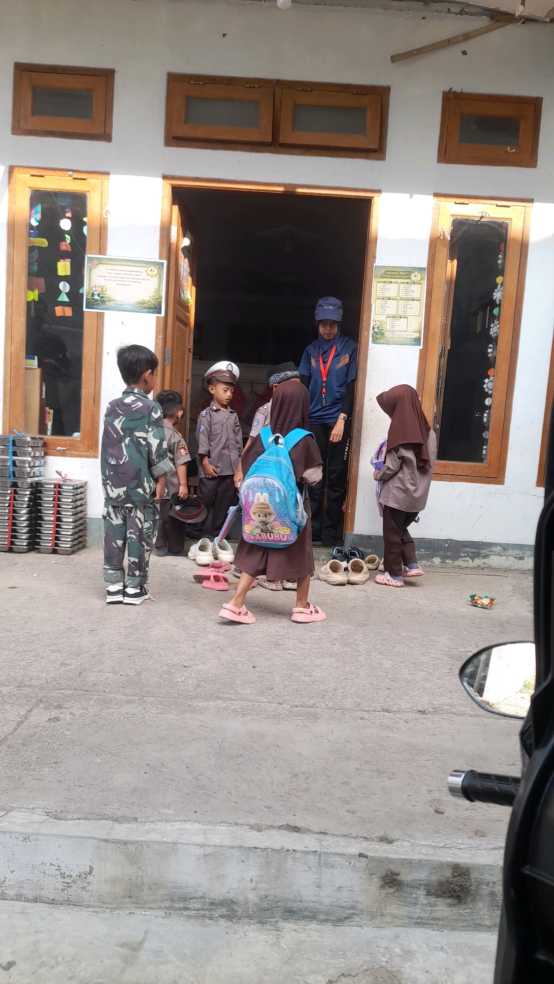 The image depicts a group of children and one adult in what appears to be a schoolyard or the entrance to a building in Selaawi, Indonesia. The building has large wooden doors and windows. In the foreground, several children are gathered. One boy is wearing a camouflage uniform. Another boy is dressed in a police-style uniform with a hat. A girl in a brown headscarf and dress is carrying a blue backpack with a character image on it. Another girl, also in a brown headscarf and dress, is standing nearby. An adult, wearing a blue uniform and a cap, is standing in the doorway. Shoes are scattered on the ground in front of the children. Stacks of metal trays are visible to the left of the entrance. The ground is paved with concrete. A motorcycle's side mirror is visible in the bottom right corner of the image.