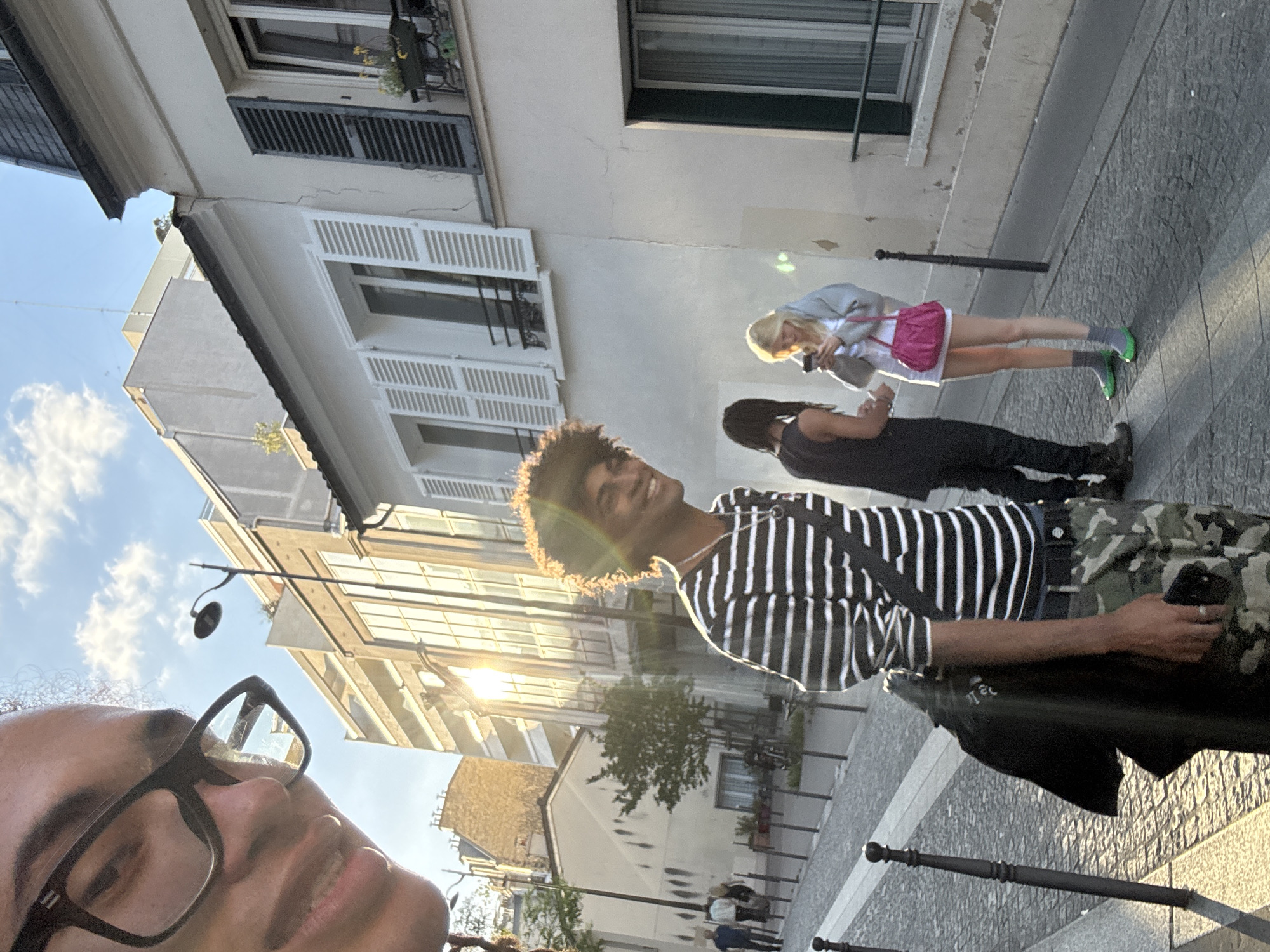 The image depicts a street scene in Paris, France. A person wearing glasses is taking a selfie, partially visible in the foreground. Behind them, another individual is smiling and wearing a striped black and white shirt and camouflage pants. Further back, two other people are standing on the sidewalk: one is wearing a gray jacket and pink bag, looking at their phone, and the other is wearing black clothing. Buildings with windows and shutters line the street. The lighting suggests it is either early morning or late afternoon, with the sun creating a flare in the image. The street is paved and features decorative metal posts along the curb.