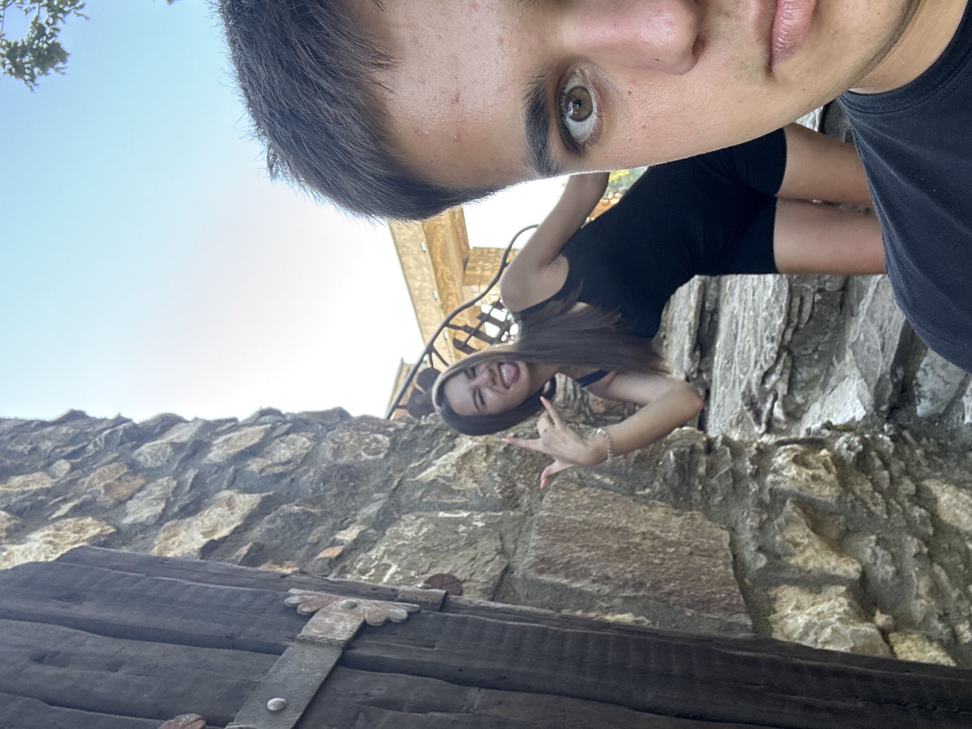 A young woman with long dark hair, wearing a black dress, makes a rock-on hand gesture and sticks out her tongue while standing on stone steps near a weathered wooden door and a stone wall. In the foreground, a young man's face is captured in a selfie, his right eye looking directly at the camera. The background shows a clear blue sky and architectural elements. This scene appears to be set within the environs of the Karla Laka Waterfall in Lukovit, Bulgaria, a natural attraction known for its stone formations and historical structures.