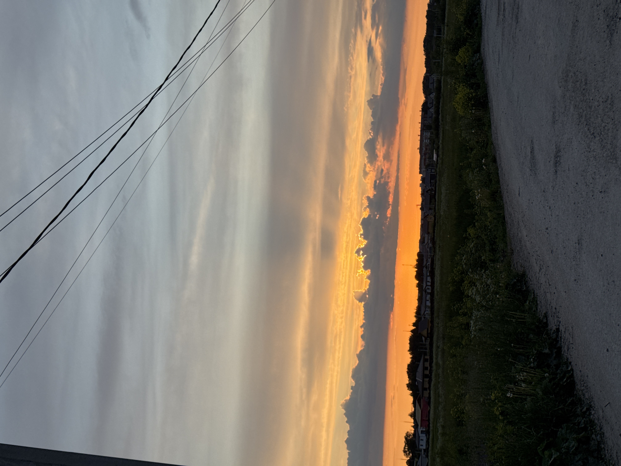 The image displays a serene landscape captured during either sunrise or sunset in a rural or semi-rural setting, likely near Горный, Russia. The sky occupies the majority of the frame, showcasing a dramatic transition of colors. On the left, the sky is a soft blue-grey, gradually warming to vibrant oranges and yellows towards the right, where the sun's low position casts a brilliant glow. Various cloud formations are present, ranging from thin, streaky wisps to denser, darker clouds that catch and filter the warm light, creating an atmospheric effect.
Several dark power or utility lines traverse the upper left quadrant of the sky, adding a subtle element of infrastructure to the natural scene. Below the dynamic sky, a strip of dark green vegetation, composed of grass and bushes, runs across the middle ground. Along the horizon line, silhouetted against the bright orange glow, are numerous small buildings or houses, suggesting a distant village or settlement. The foreground on the right features a wide, light-colored gravel or dirt road that appears to stretch into the distance. No individuals or animals are visible, and there is no discernible text within the image. The overall impression is one of tranquility and natural beauty at the cusp of day or night.