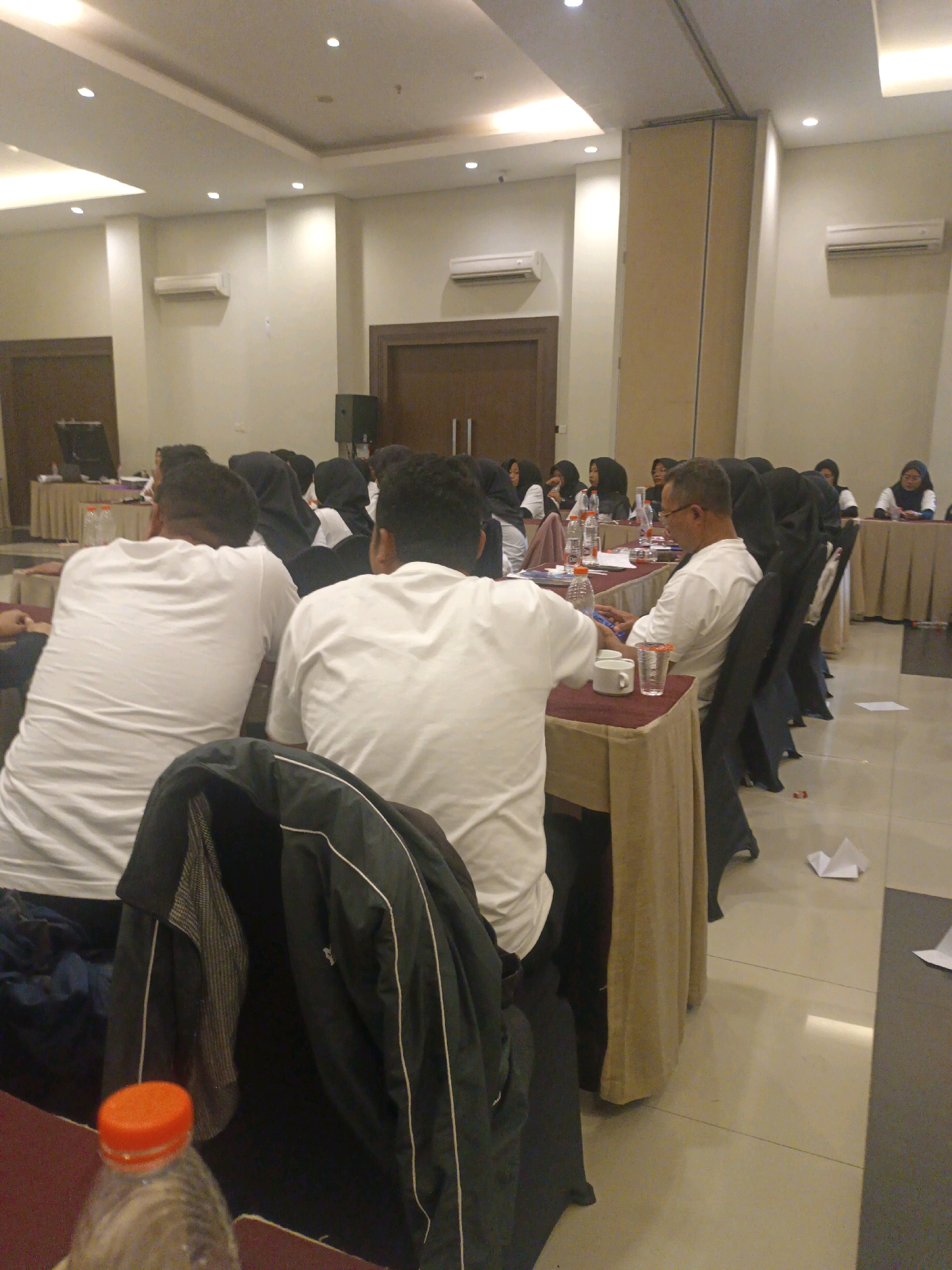 The image depicts an indoor event, likely a seminar or conference, taking place in a room with a tiled floor and white walls. The room is equipped with multiple air conditioning units and recessed lighting. In the foreground, two individuals are seated, facing away from the camera, both wearing white t-shirts. A dark jacket is draped over a chair in front of them, and a plastic water bottle with an orange cap is visible. Further into the room, a long table is set up with chairs, where more people are seated. Some individuals, particularly women, are wearing headscarves. Items on the table include water bottles, cups, and what appear to be laptops. The overall atmosphere suggests a formal gathering. The location is identified as Jogoloyo, Indonesia.