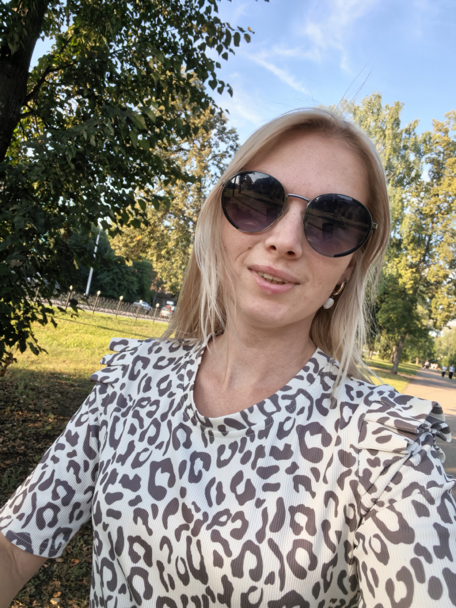 A woman with blonde hair, wearing sunglasses and a leopard print top, poses for a selfie in an outdoor setting. The background suggests a park or city street with trees and a pathway. The lighting indicates it is daytime. The location cannot be determined from the image itself and no identifying features of Sterlitamak, Russia are present.