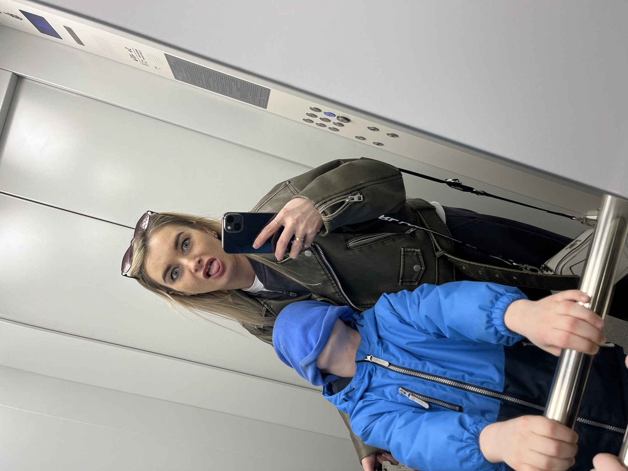 A woman with long blonde hair and wearing a green leather jacket is taking a selfie with a child in a blue jacket. The child is holding onto a metal bar. They appear to be inside an elevator. The control panel of the elevator is visible in the upper portion of the image. This photograph was possibly taken in the city of Chaikovsky, Russia.