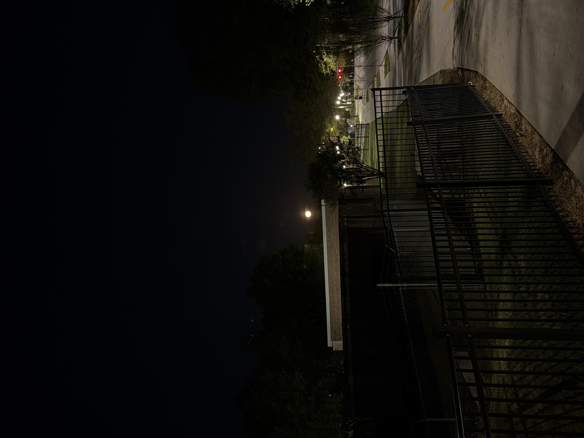 A nighttime photograph shows a dark urban scene, likely in a city. The presence of streetlights and a traffic signal suggests a municipal setting. A dark chain-link fence runs along the right side of the image, separating the viewer from what appears to be a park or green space. There are structures with flat roofs and some lights. In the distance, a bright object that looks like the moon can be seen. The general characteristics of the scene, like the lighting style of the lamp posts, are similar to those in Atlanta, United States of America.