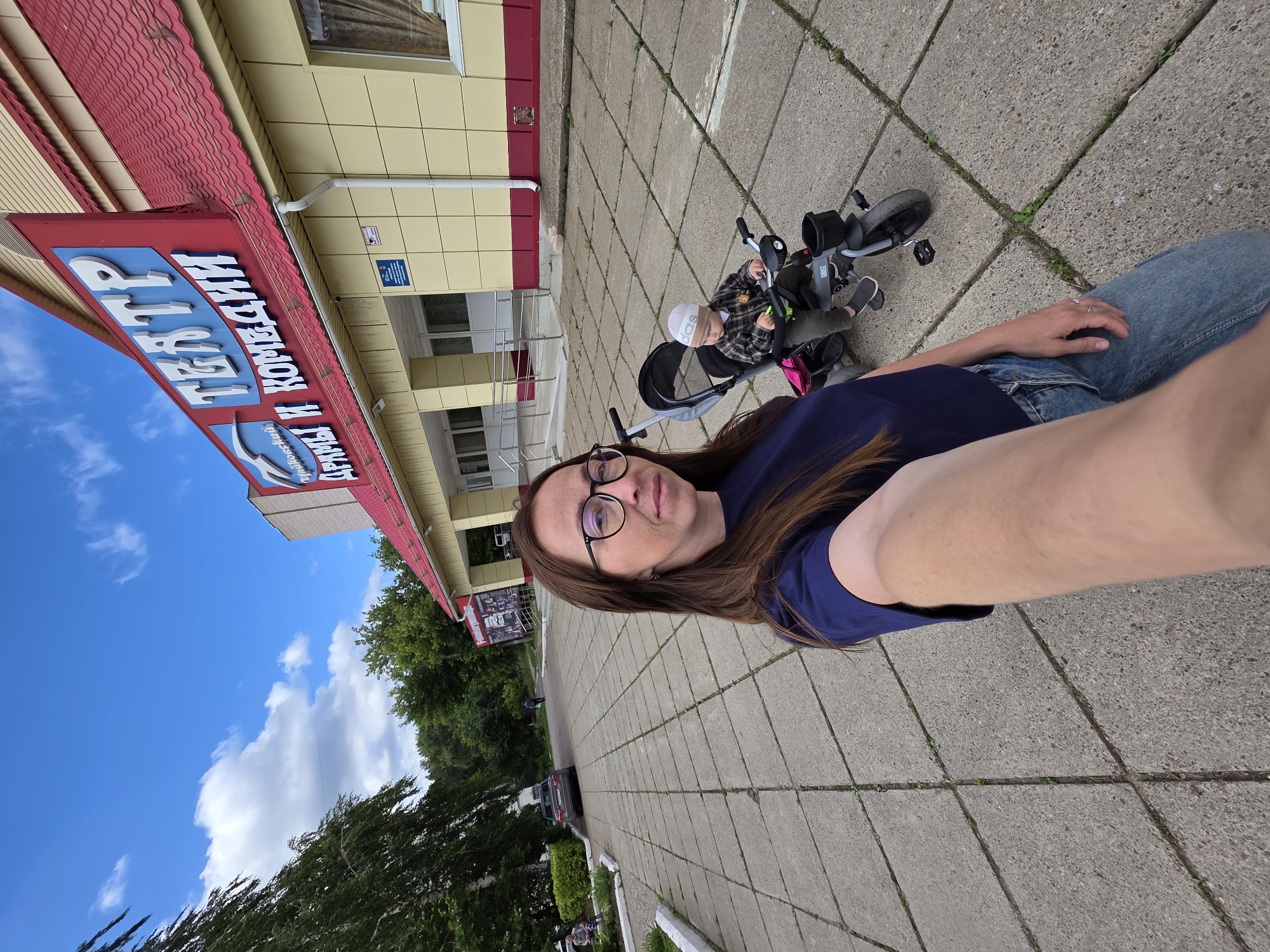 A woman wearing glasses and a dark blue shirt takes a selfie outside a theater. Next to her, a toddler in a plaid shirt and white hat rides a tricycle. The theater, identified by signs as "ТЕАТР" (Theater) and "ДРАМЫ И КОМЕДИИ" (Drama and Comedy), has a yellow facade with red trim and a red tiled roof. The ground is paved with square concrete tiles. In the background, trees and a car are visible. This scene is in the city of Chaikovsky, Russia.