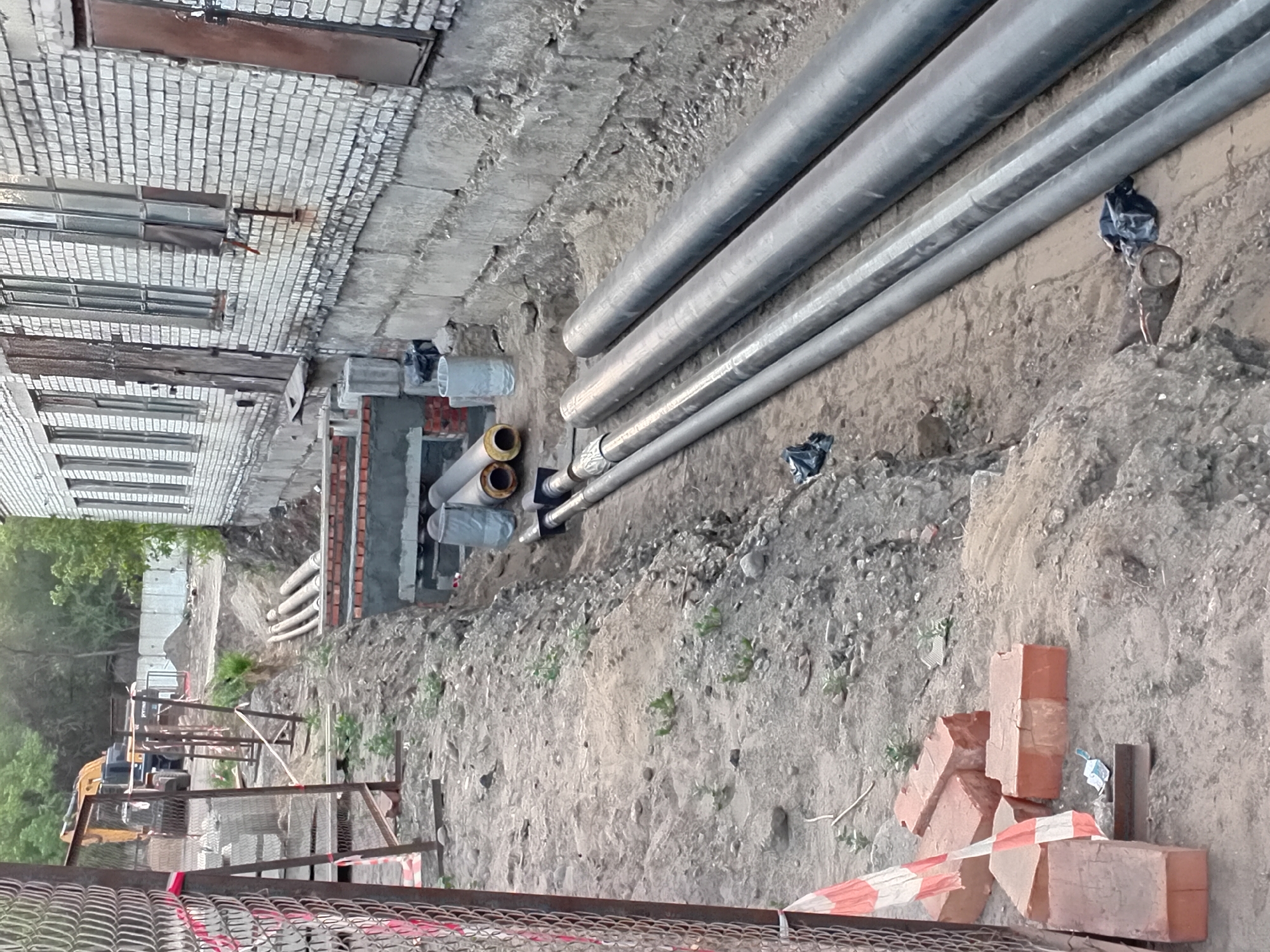 The image depicts an outdoor construction site in Semey, Kazakhstan. The scene features a trench that has been dug alongside a brick building. Several large pipes are laid out, some in the trench and others on the ground next to it. These pipes appear to be part of an underground utility or construction project. In the background, an excavator is visible, indicating ongoing construction work. The ground is a mixture of dirt, sand, and some debris, with some vegetation growing in patches. There are also stacks of bricks and construction materials scattered around the area, suggesting preparation for further building or repair. The overall impression is one of an active construction zone.