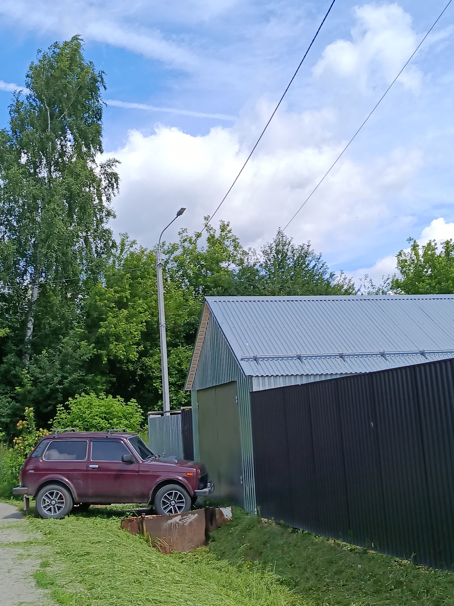 A maroon SUV is parked on a grassy area next to a dark, corrugated metal fence. Behind the fence is a building with a silver, corrugated metal roof. A street light on a pole stands to the left of the building, amidst a backdrop of lush green trees and a blue sky with scattered clouds. The scene appears to be in a rural or suburban setting, possibly in the city of Vereya, Russia, given the context of the prompt.