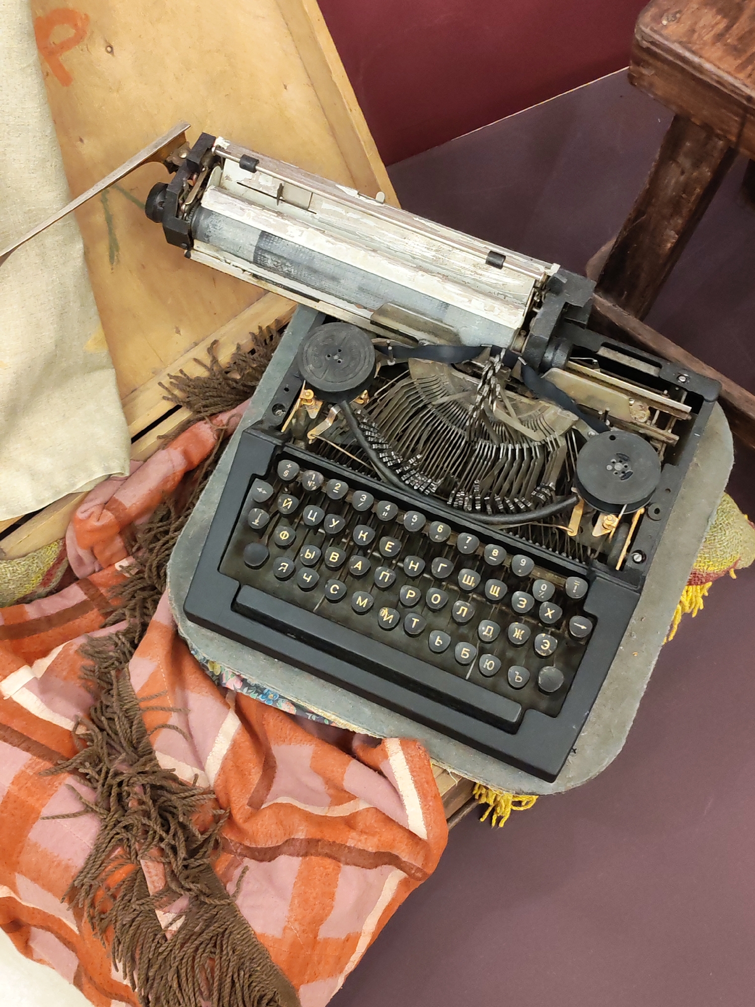 An overhead shot captures a vintage, black typewriter with a Cyrillic keyboard, prominently displayed indoors in Izhevsk, Russia. The typewriter's metal typebars are exposed, and black ribbon spools are visible. Its silver-toned carriage sits above the keyboard.
The typewriter rests on a textured, possibly padded, grey surface, which in turn sits on a wooden crate or box. A plaid blanket or scarf, featuring a large orange and pink/light-colored check pattern with brown fringes, is draped around the left side of the typewriter, partially covering the wooden surface. A light-colored fabric, possibly canvas, is also visible on the wooden surface in the upper left.
In the background, a dark red or maroon surface (likely a floor or wall) provides a muted backdrop, with part of a wooden stool or chair leg visible on the right. The scene appears to be a still-life arrangement or a display, evoking a sense of nostalgia.
Visible text includes numerous Cyrillic letters on the typewriter keys such as "Й", "Ц", "У", "К", "Е", "Н", "Г", "Ш", "Щ", "З", "Х", "Ф", "Ы", "В", "А", "П", "Р", "О", "Л", "Д", "Ж", "Э", "Я", "Ч", "С", "М", "И", "Т", "Ь", "Б", "Ю", and "Ъ", alongside numbers and some symbols. An orange painted letter "P" is also faintly visible on the wooden surface in the upper left. The lighting suggests an indoor setting, with no specific time of day discernible.