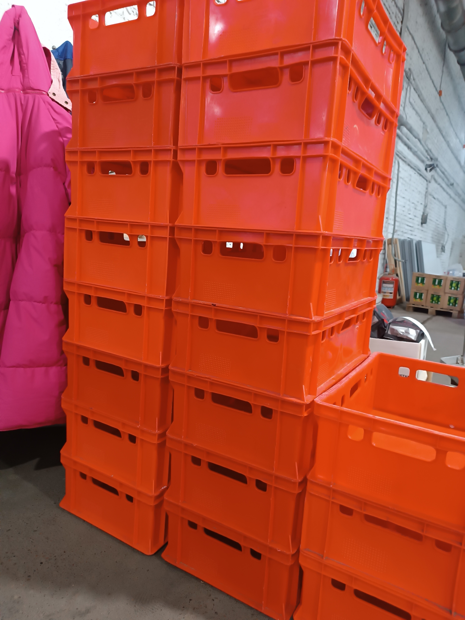 The image shows a stack of orange plastic crates in what appears to be an indoor storage area. A pink jacket is partially visible on the left. In the background are other items, including a fire extinguisher and stacked goods on a pallet. The location is identified as Chaikovsky, Russia. The setting is possibly a warehouse or a storage facility. There are no people visible in the image.
