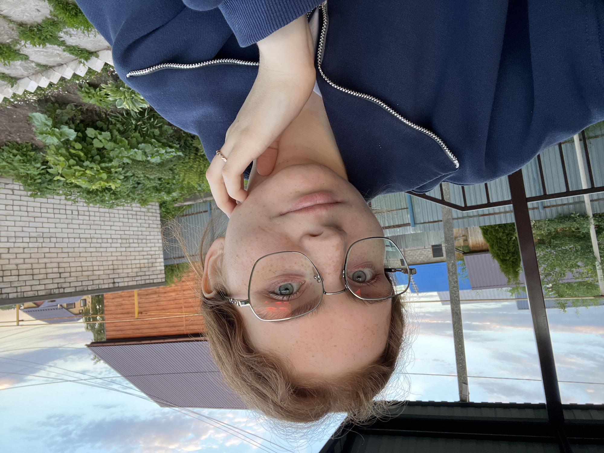 A young woman is depicted in a close-up, headshot-style image, appearing to be a selfie. She has light brown hair, which is slightly disheveled around her face, and numerous freckles are visible across her nose, cheeks, and forehead. She wears large, rectangular-framed eyeglasses, through which her light-colored eyes are visible, looking directly at the viewer. Her lips are slightly parted.
She is dressed in a dark blue zipped jacket or hoodie, with the silver zipper prominent on the front. Her right hand, adorned with a simple golden ring on her ring finger, is resting near her neck, with her left hand likely beneath it, out of view.
The background suggests an outdoor, possibly suburban or rural setting. Directly behind her, to the viewer's left, is a manicured garden area with various green plants and a pathway bordered by white stones. Further back, a light-colored brick wall and a wooden fence are visible. To the right, various building structures with corrugated metal roofs (brown and blue) are seen, alongside utility poles and scattered trees and greenery. The sky, featuring soft clouds and hues, indicates late afternoon or early evening. The overall location is Novaya Tura, Russia. No visible text is present in the image.