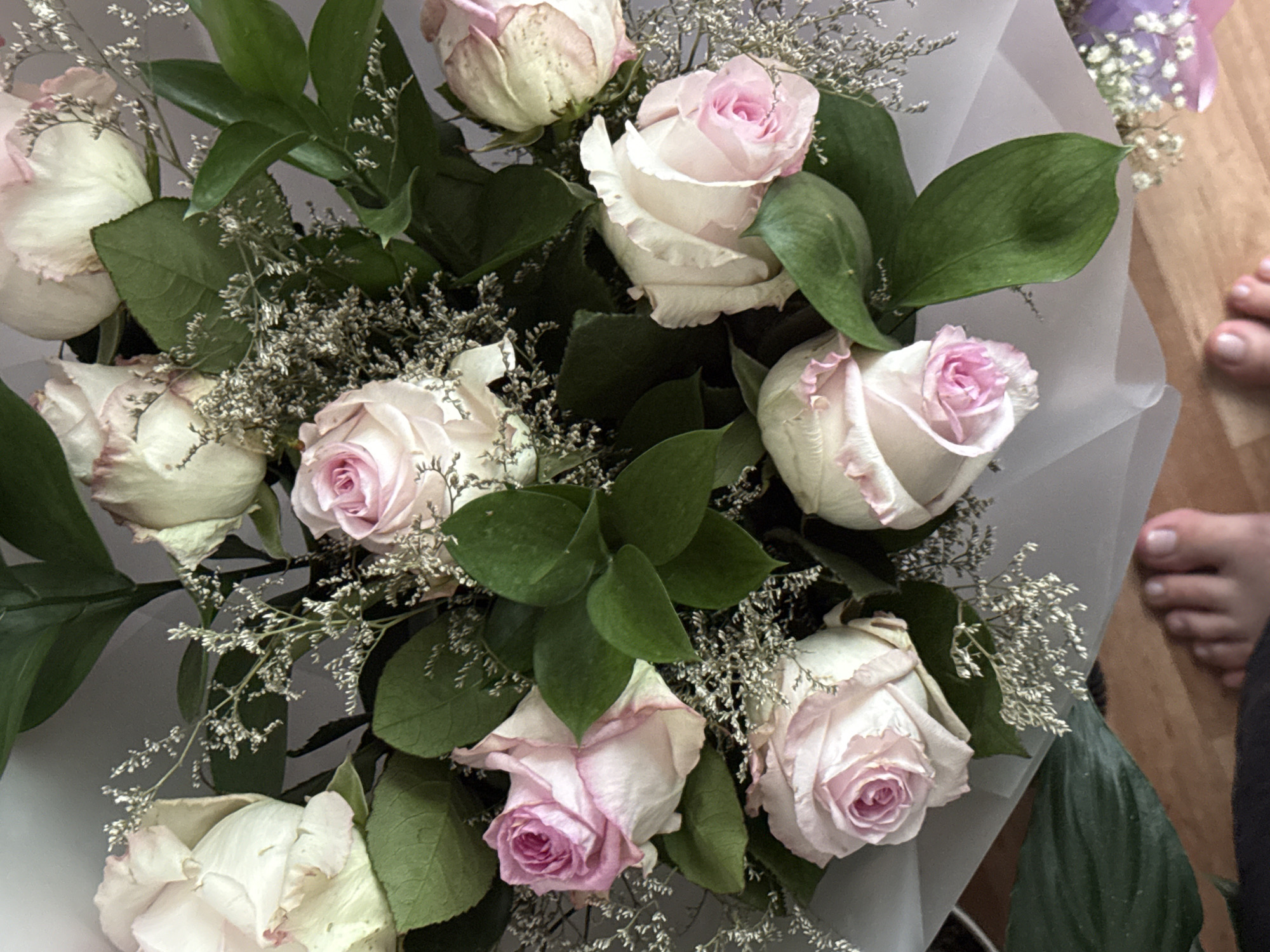 The image shows a bouquet of roses with white and pink petals, interspersed with greenery and small white flowers. The bouquet is wrapped in a light-colored material. Part of a human foot with visible toes is in the lower right corner of the image. No information indicates this photo was taken in Chaikovsky, Russia.